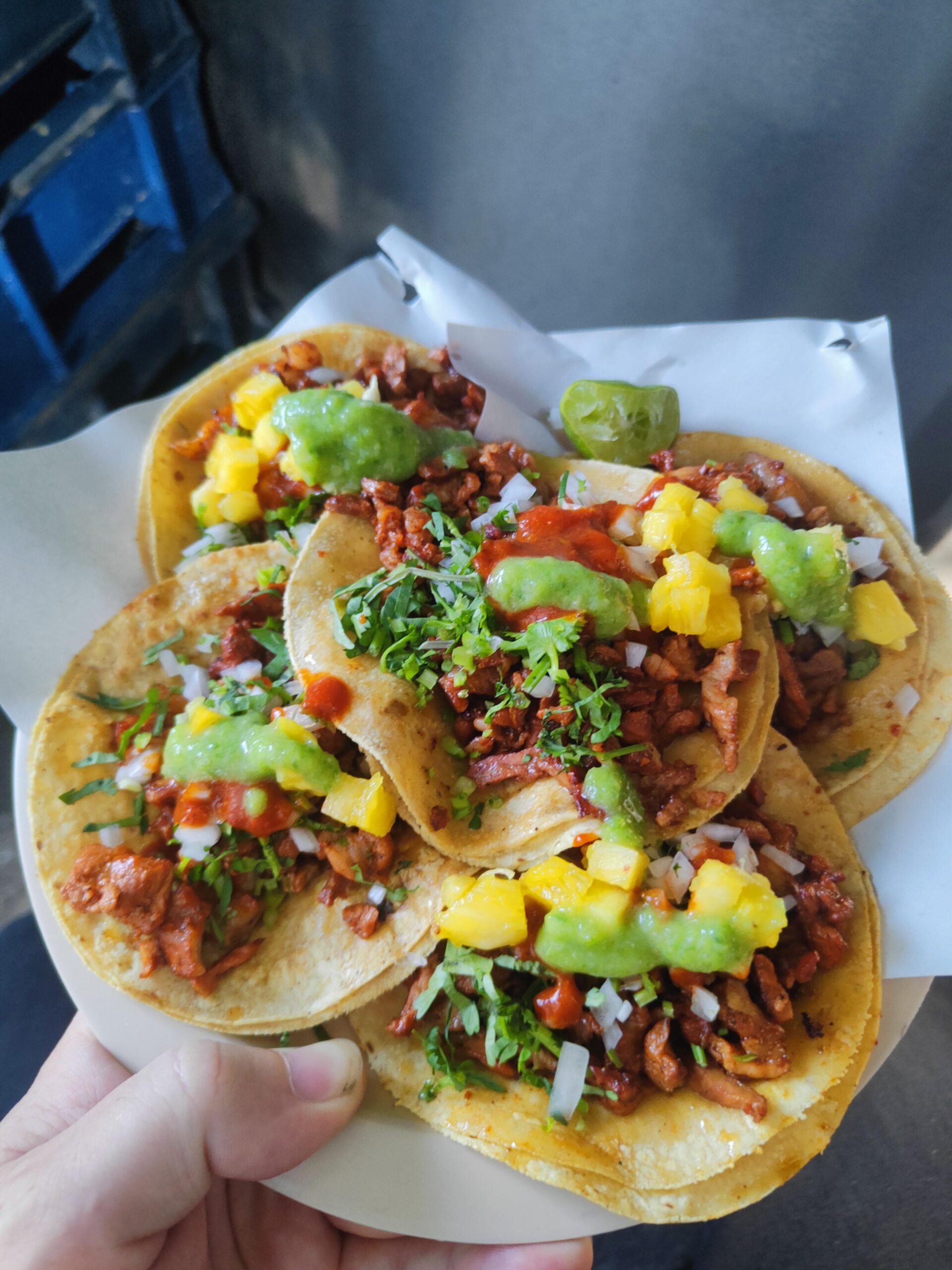 A hand holds a plate with four tacos filled with seasoned meat, chopped onions, cilantro, pineapple chunks, and topped with green salsa. The tacos are served on soft corn tortillas.