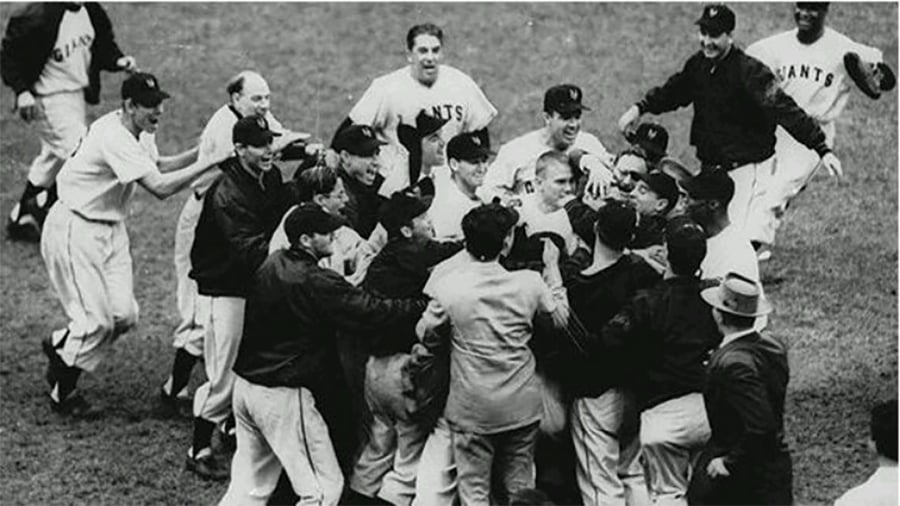 A group of baseball players and coaches, wearing "Giants" uniforms, enthusiastically celebrate together on a field, surrounding a central figure in a moment of excitement and joy.