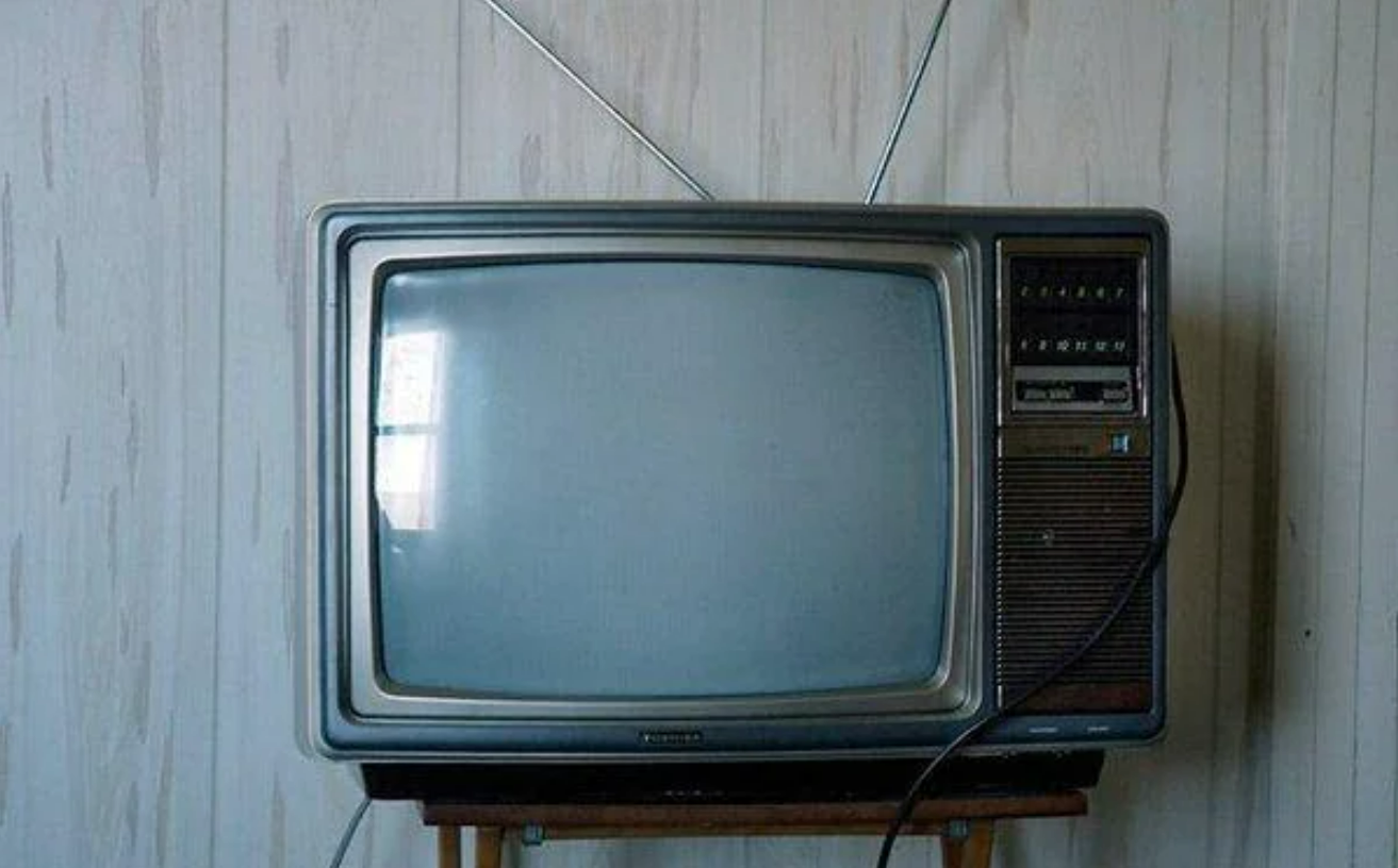 A vintage box-style television with a rounded screen sits on a small table. It has two antennae sticking up and buttons on the right side. The TV is against a light-colored, striped wall.