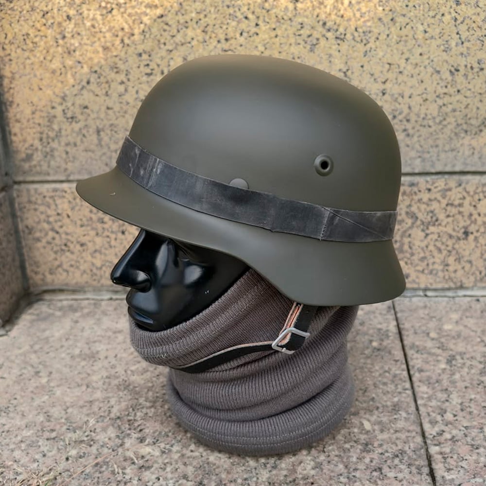 A matte green military-style helmet with a black strap is displayed on a black mannequin head, which is wrapped in a gray scarf. The background consists of speckled beige tiles.