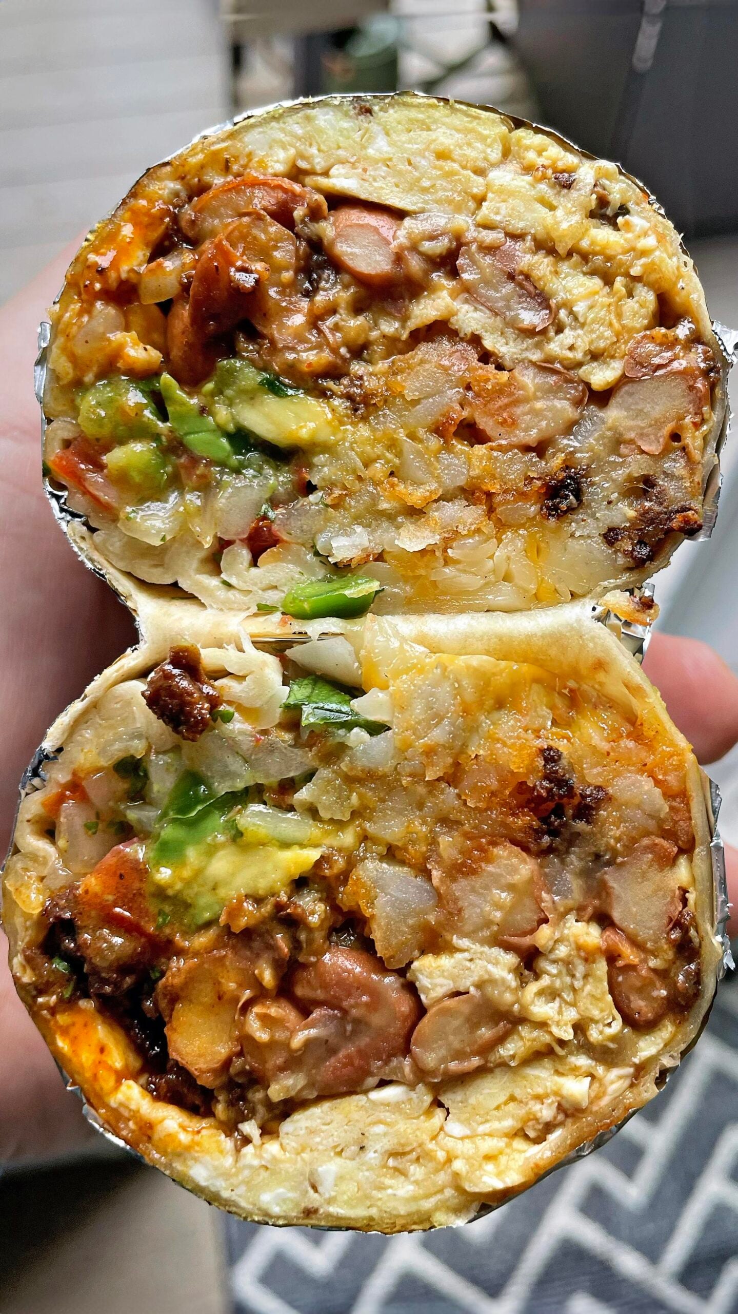 A close-up of a burrito cut in half, showing layers of rice, beans, avocado, potatoes, eggs, salsa, and cheese wrapped in a flour tortilla with foil around the edges. A hand is holding the burrito halves.