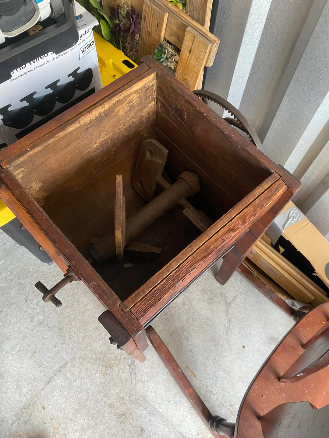 A wooden, vintage butter churn with an open lid reveals internal wooden paddles and a crank mechanism. The churn is sitting on a concrete floor, surrounded by storage items and a yellow bin.