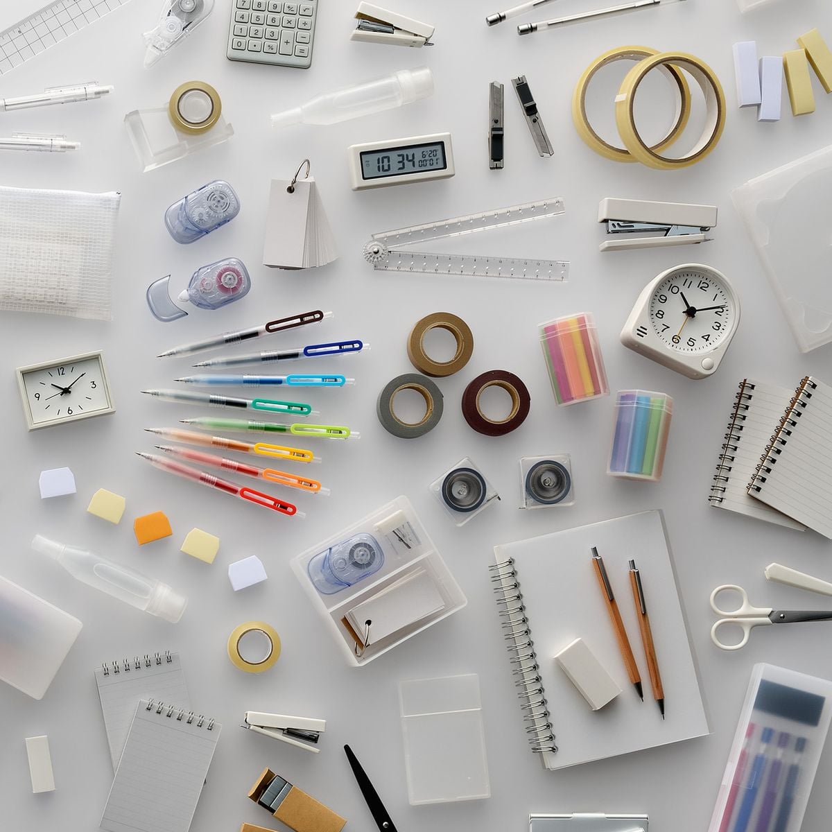 Assorted office and stationery supplies, including pens, notebooks, tape, sticky notes, scissors, rulers, clocks, and containers, arranged neatly on a white surface.