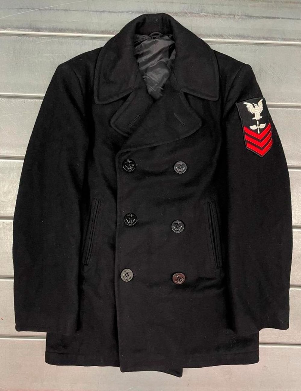 A black double-breasted military peacoat with six buttons, wide lapels, and a patch featuring a white eagle and red chevrons on the left sleeve, displayed on a wooden surface.