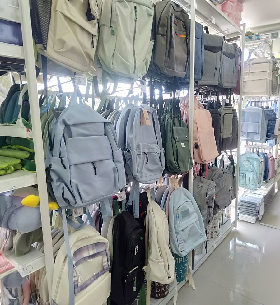 Rows of colorful backpacks hanging on display racks in a well-lit store, with shelves above and below holding additional bags and various school supplies.