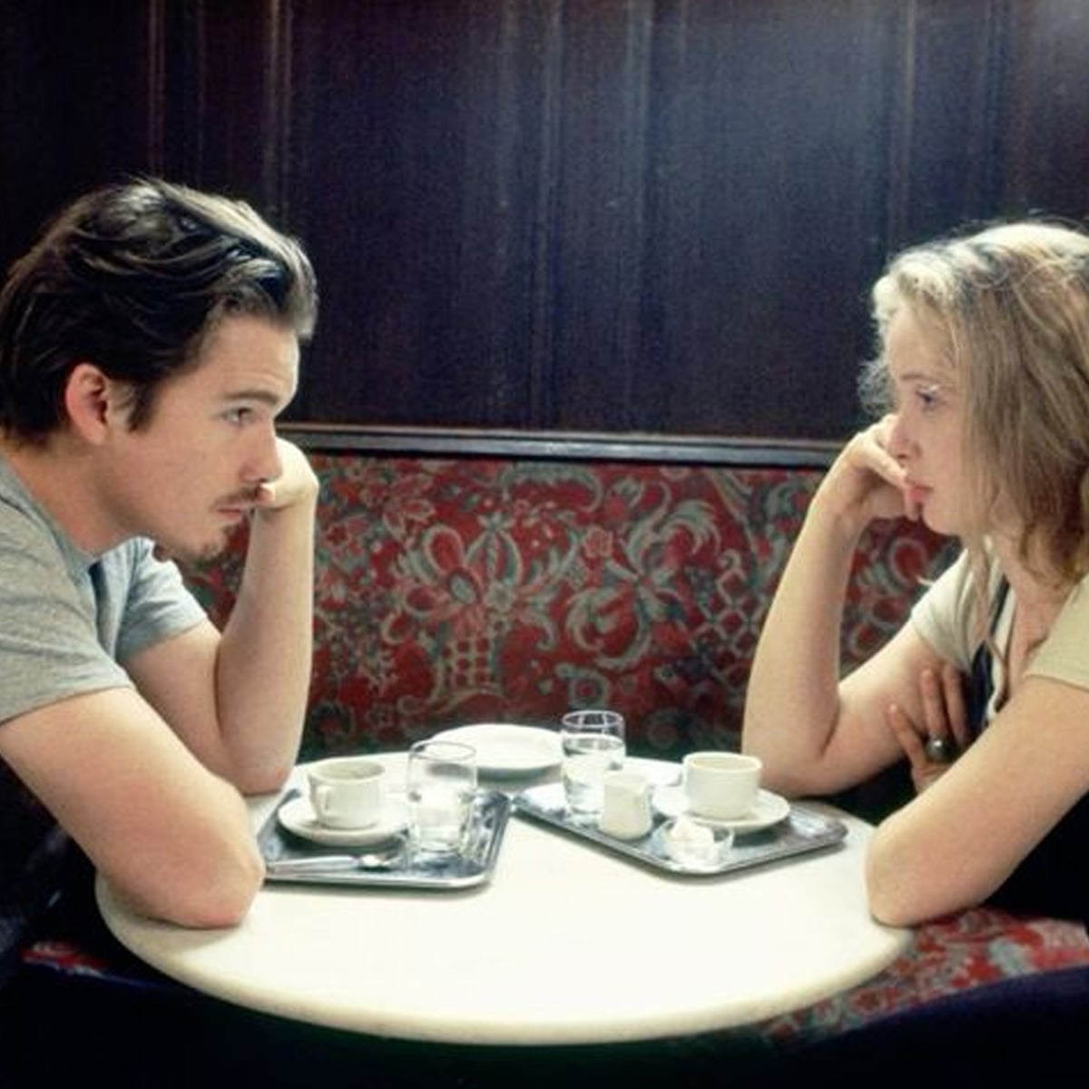 A man and a woman sit across from each other at a small round table in a cafe, leaning in with serious expressions, with coffee cups, glasses, and trays in front of them.