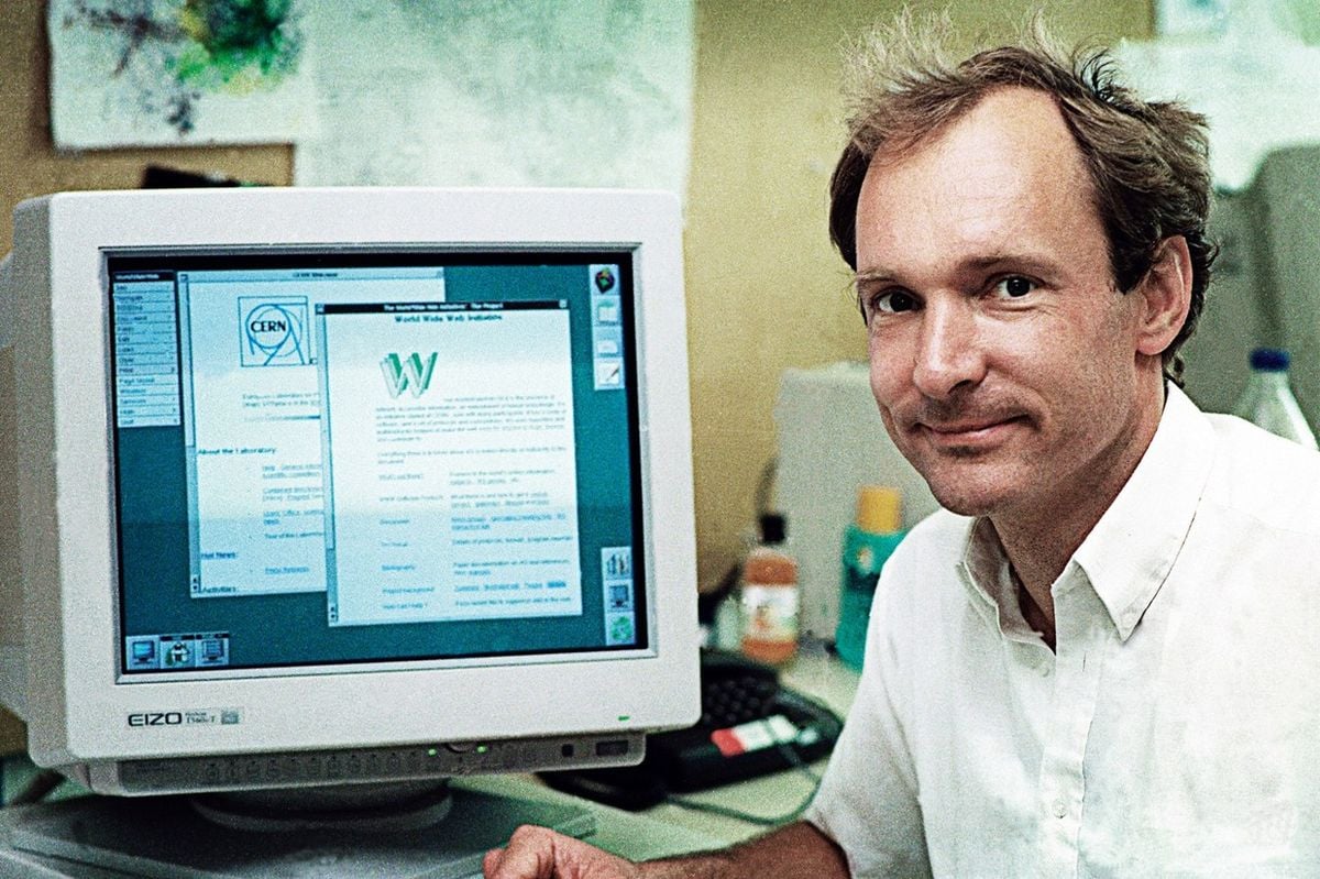 A man in a white shirt sits at a desk beside an old computer monitor displaying an early web page. The background shows a cluttered office with papers and electronics.