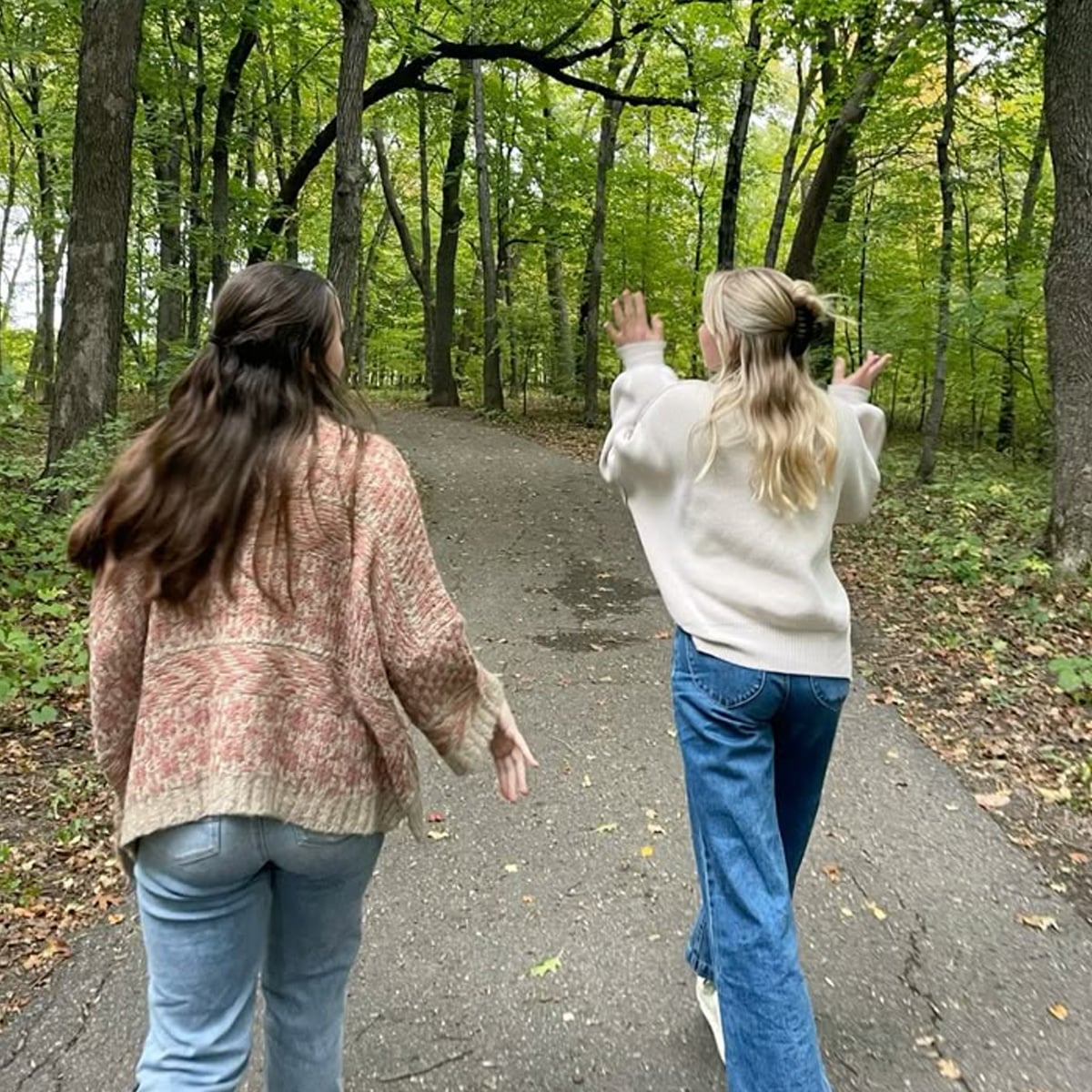 Two people with long hair walk on a paved path through a green, wooded area. Both wear sweaters and jeans. One gestures with raised hands while the other walks beside her, surrounded by tall trees and leafy ground.