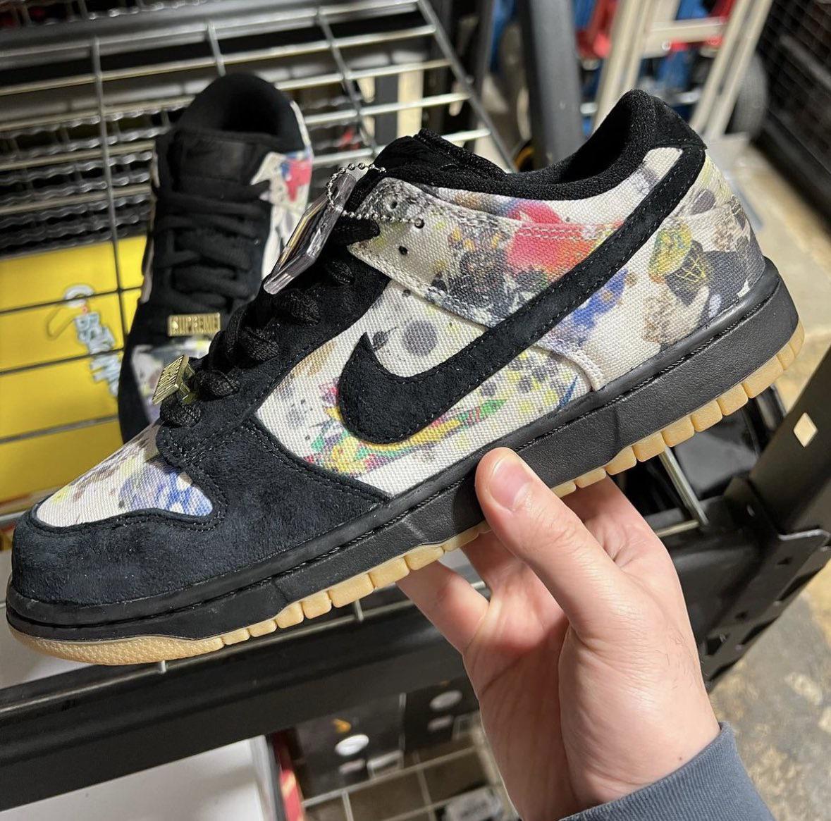 A hand holds a Nike sneaker featuring a black swoosh, black suede accents, and a multicolored abstract pattern on the upper. The shoe has a gum sole and is displayed in front of a shelving unit with another sneaker in the background.