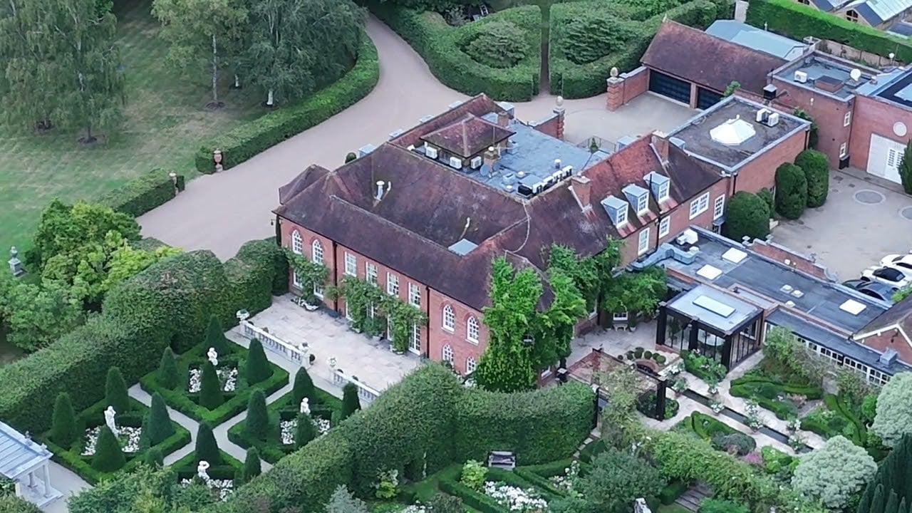 Aerial view of a large red-brick mansion surrounded by lush gardens, manicured hedges, statues, and paved walkways, with a driveway and outbuildings nearby.