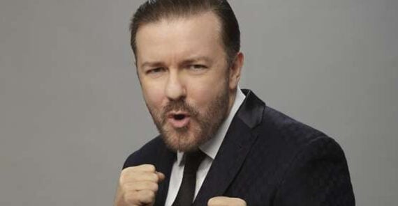 A man in a suit with neatly styled hair and a trimmed beard poses with his fists raised playfully, as if ready to box, against a plain gray background.