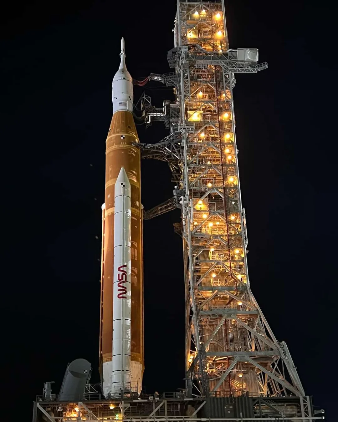 A large NASA rocket, illuminated and standing upright on a launch pad structure at night, with the tower's lights glowing against the dark sky.