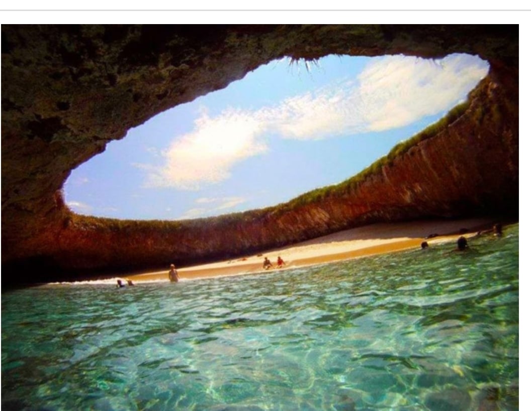 A view from inside a cave with a large circular opening above, revealing blue sky and clouds; clear turquoise water and a sandy beach with people swimming and relaxing.