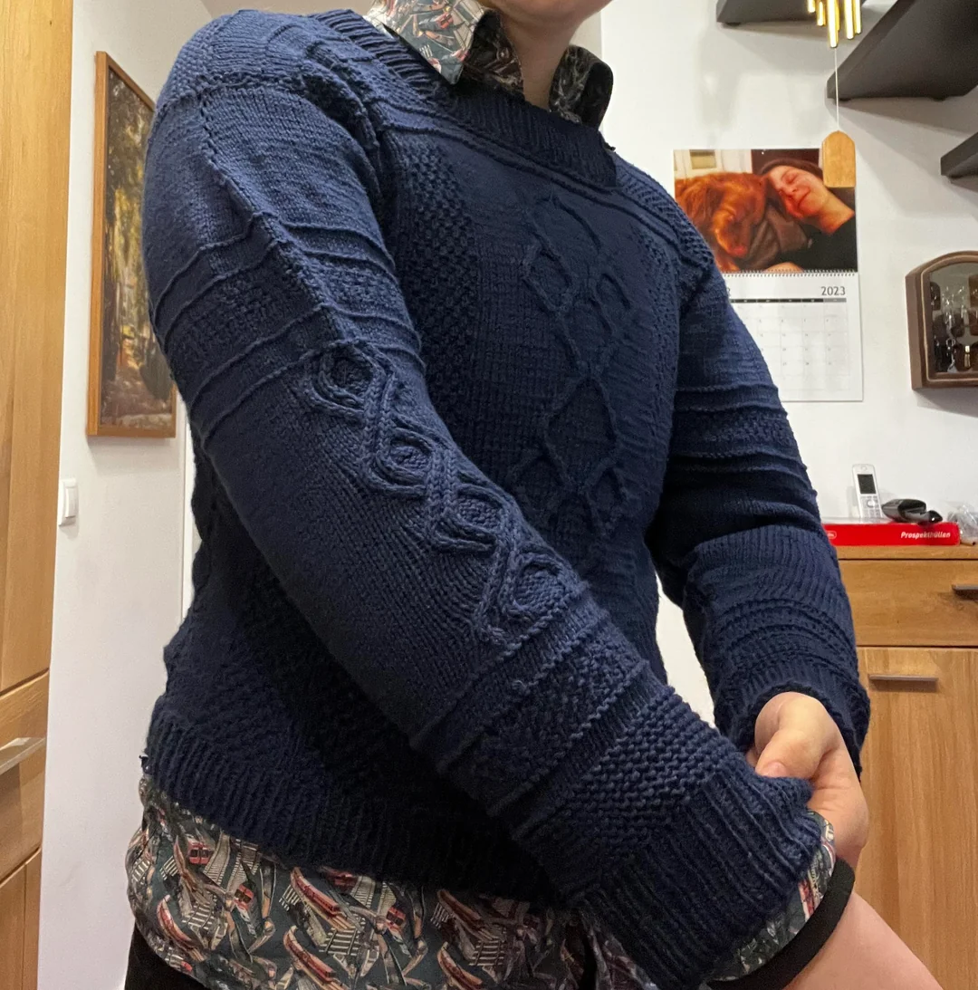 A person wearing a textured navy blue sweater over a patterned shirt is rolling up their sleeve in a kitchen. A calendar and some kitchen items are visible in the background.