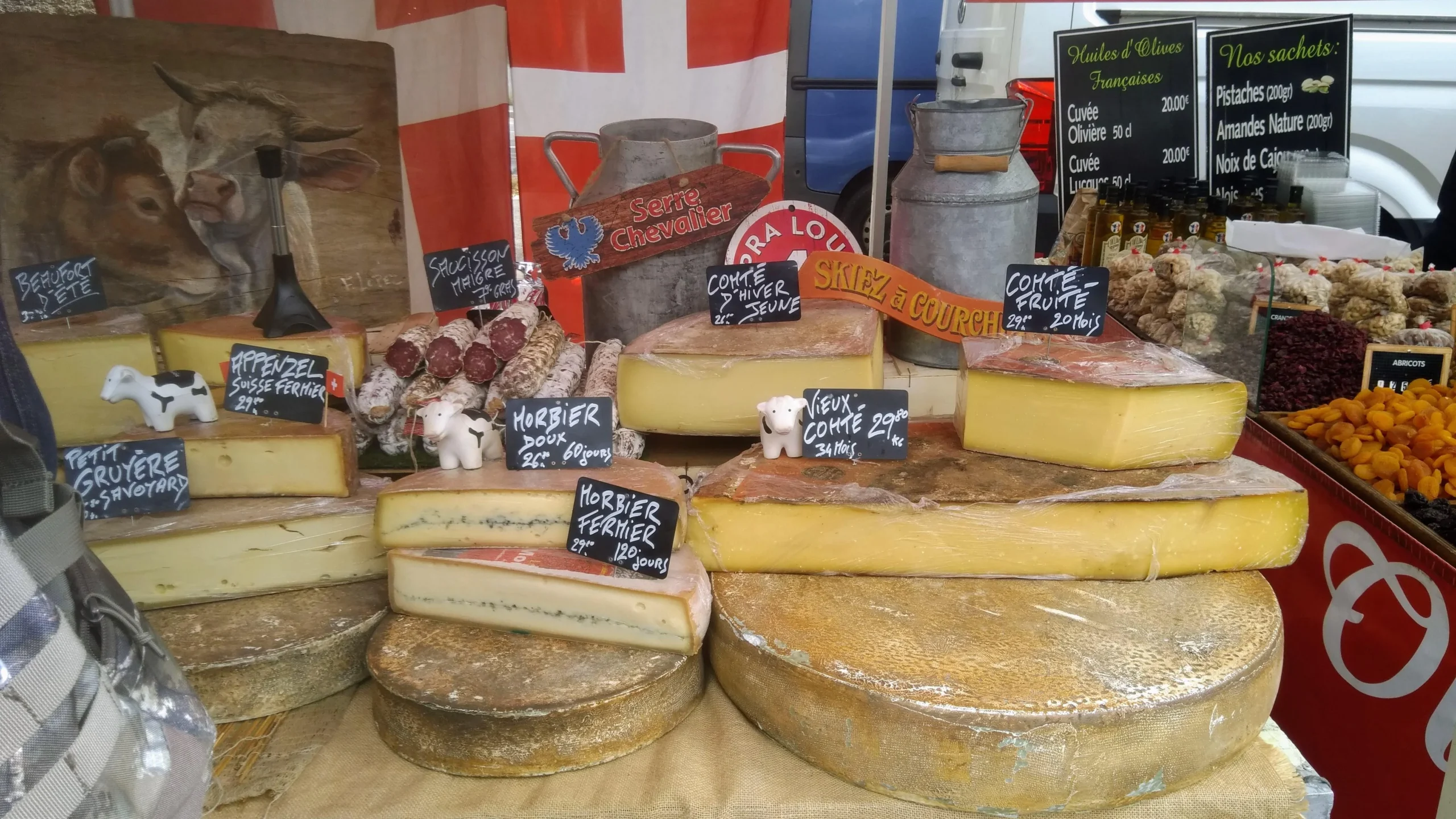 A market stall displays large wheels and wedges of cheese, some labeled Morbier and Comté, with small decorative cows, sausages, dried fruits, and a red-and-white flag in the background.