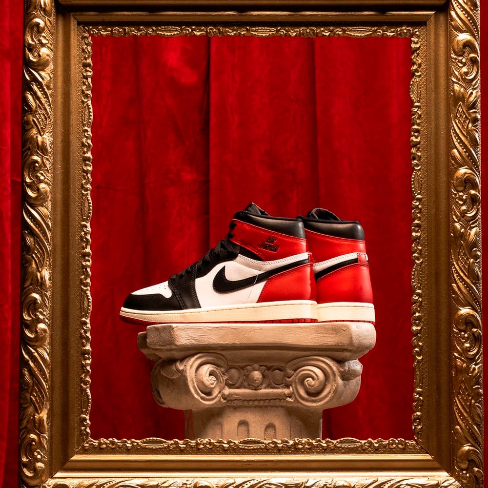 A pair of red, white, and black Nike Air Jordan sneakers displayed on a decorative pedestal, framed by an ornate gold frame, with a red velvet curtain in the background.