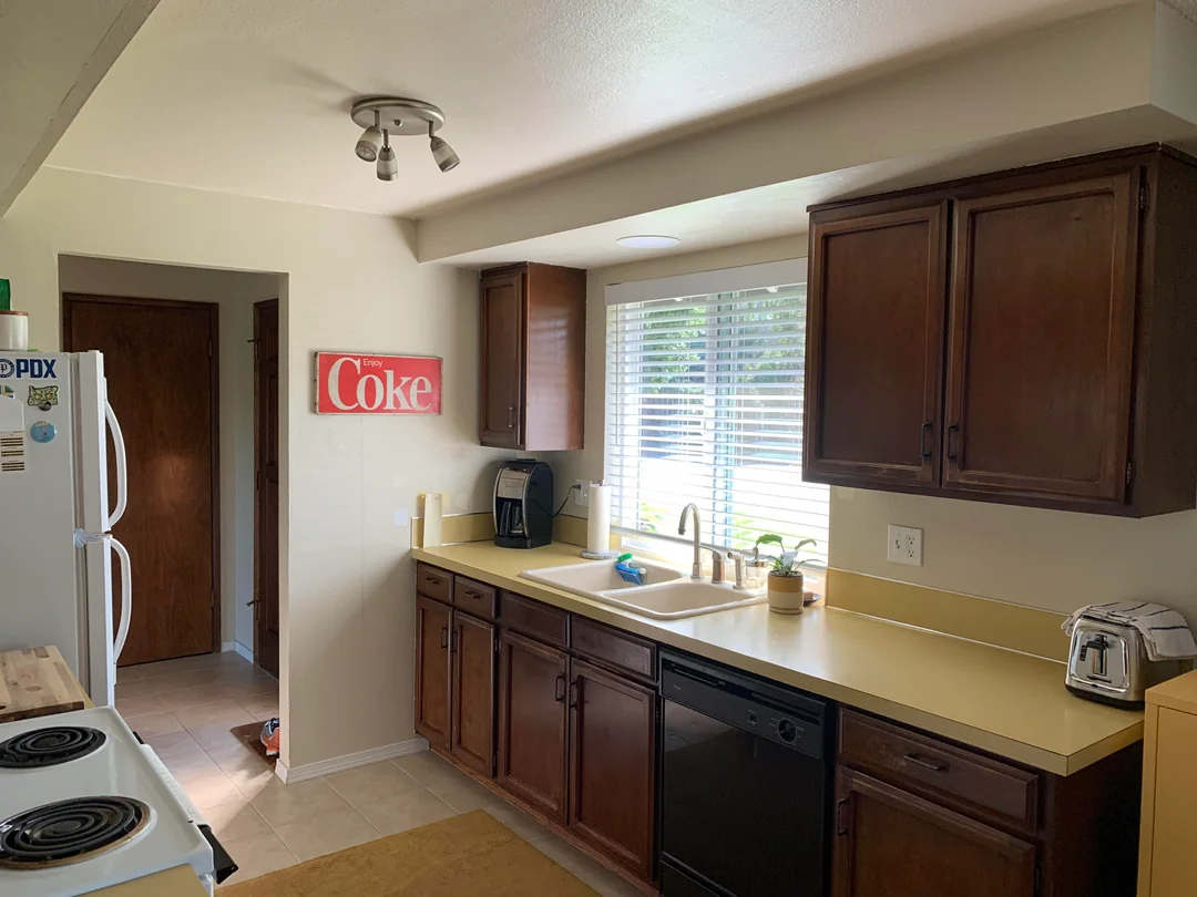 A kitchen with dark wooden cabinets, yellow countertops, a white sink under a window with blinds, and appliances including a dishwasher, toaster, coffee maker, and stove. A red Coca-Cola sign hangs on the wall.