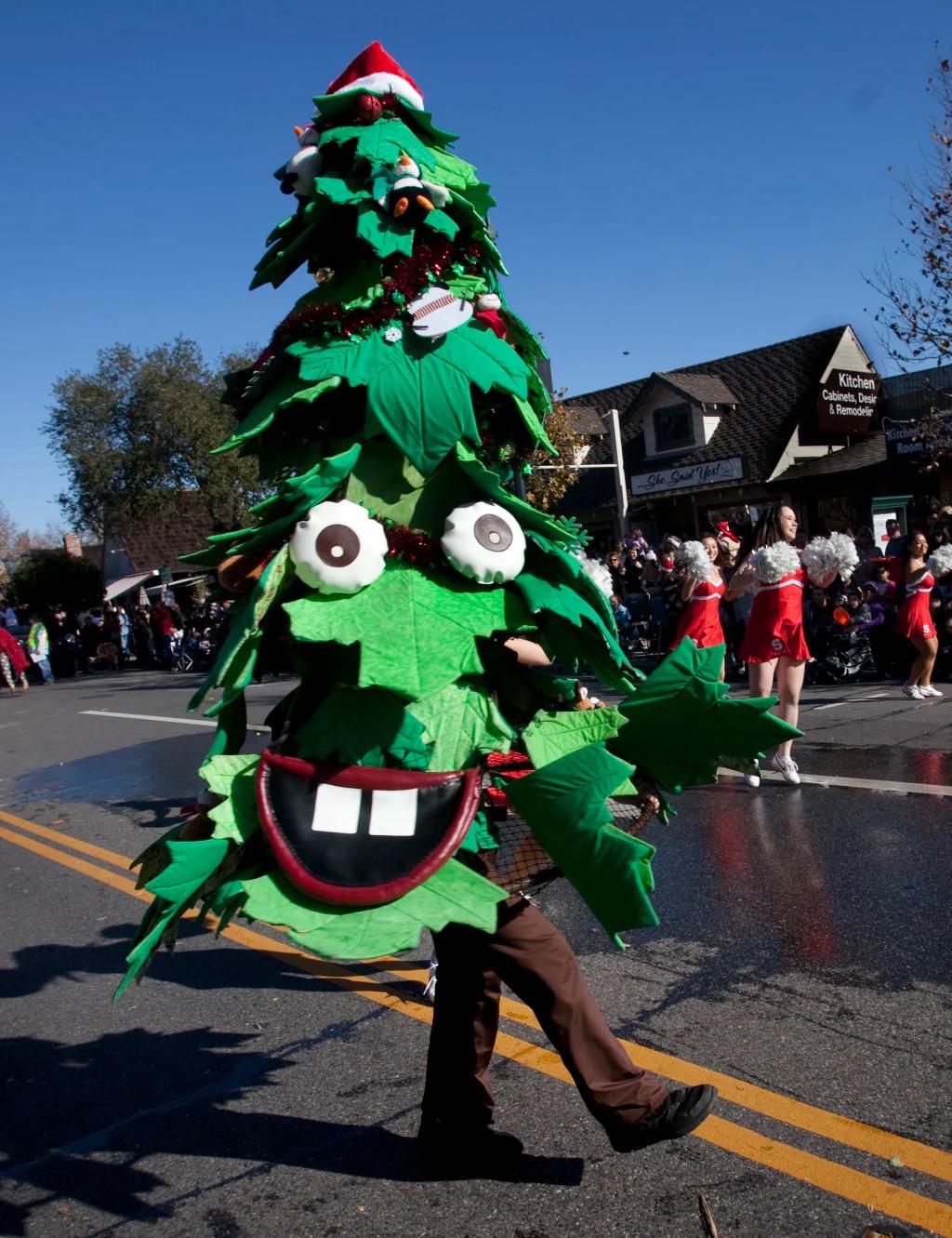 A person in a large Christmas tree costume with cartoonish eyes and a big smile walks in a parade on a sunny street, with people and shops in the background.