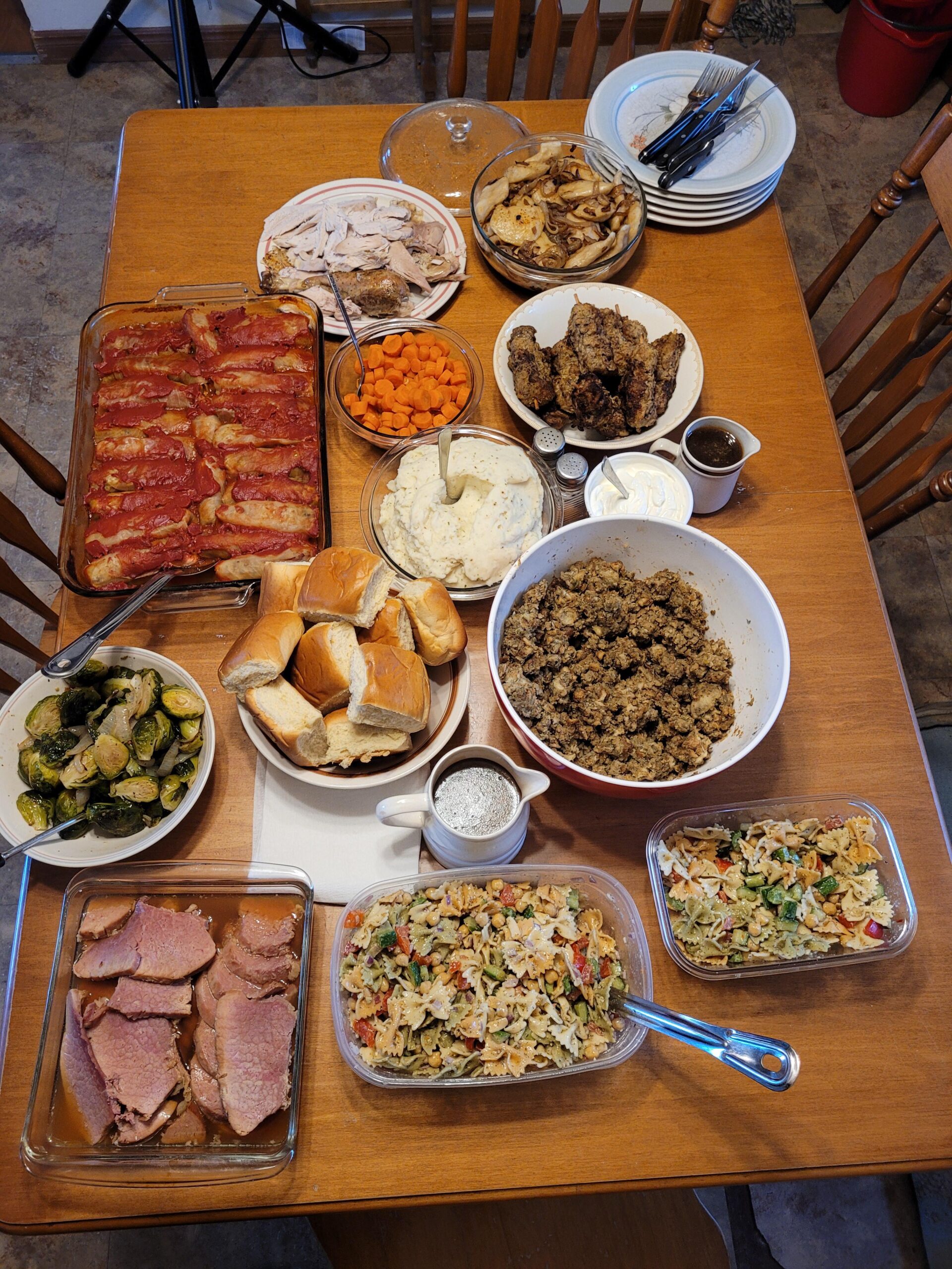 A wooden table set with various dishes: sliced turkey, ham, meatloaf, brussels sprouts, carrots, mashed potatoes, stuffing, rolls, pasta salad, roasted potatoes, fried chicken, gravy, and plates with utensils.