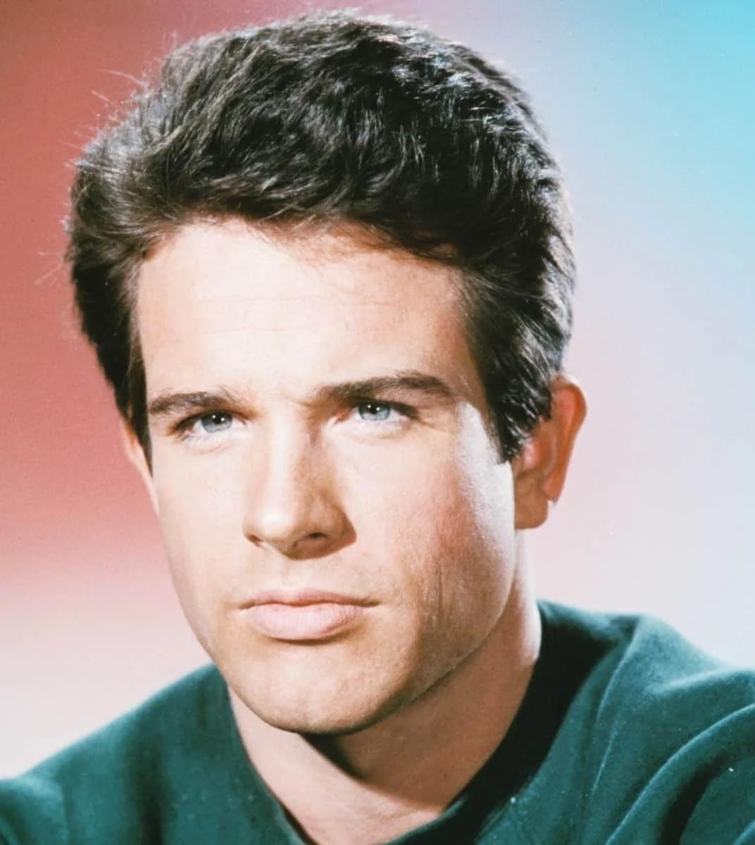 A young man with short dark hair and blue eyes stares seriously at the camera, wearing a dark green shirt against a gradient background of red and blue tones.