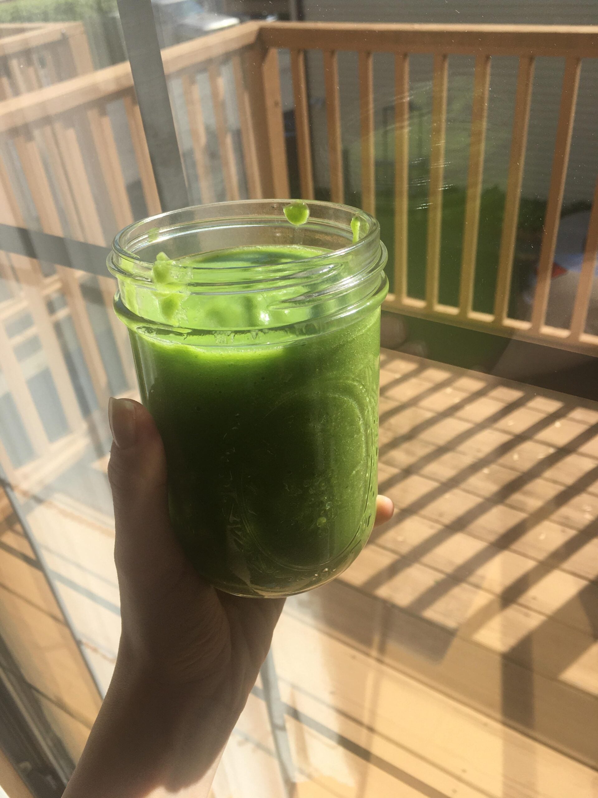 A hand holds a mason jar filled with bright green smoothie in sunlight, with a wooden deck and railing in the background.
