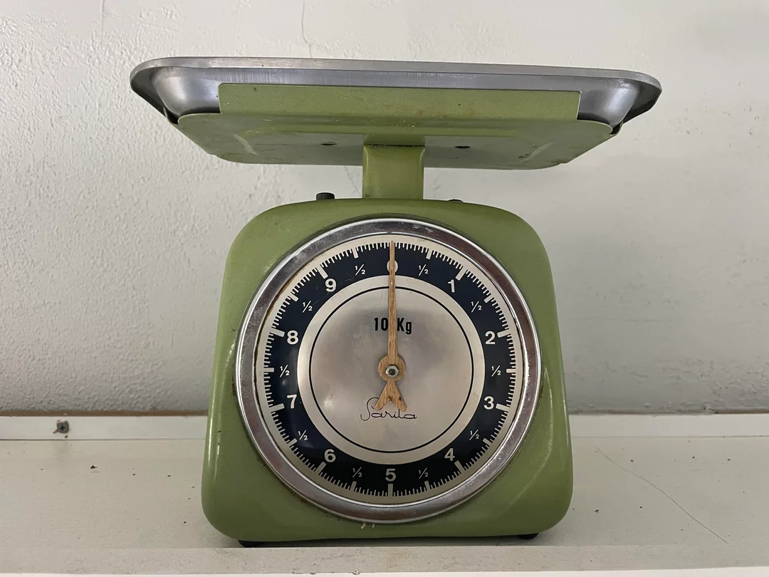 A green analog kitchen scale with a round dial and black markings, showing a maximum capacity of 10 kg, sits on a white surface against a light-colored wall. The needle points near zero.