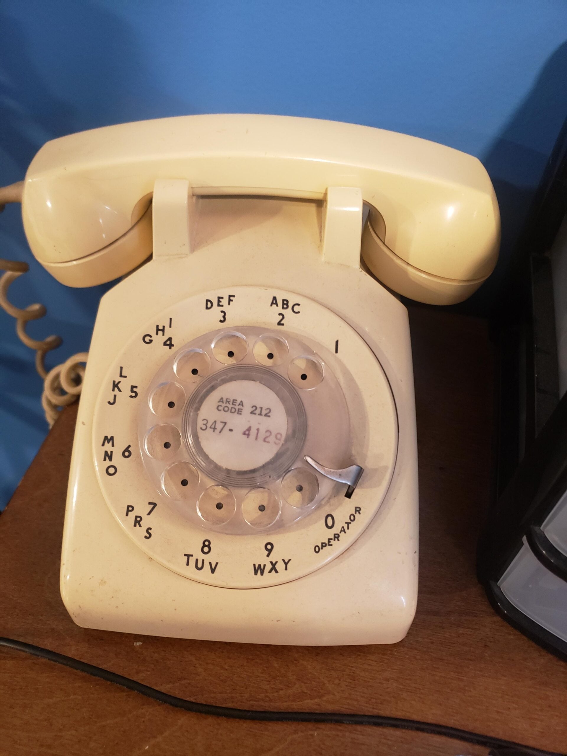 A beige rotary dial telephone sits on a wooden surface against a blue wall. The dial has black numbers and letters, and a small paper with a phone number is visible in the center.