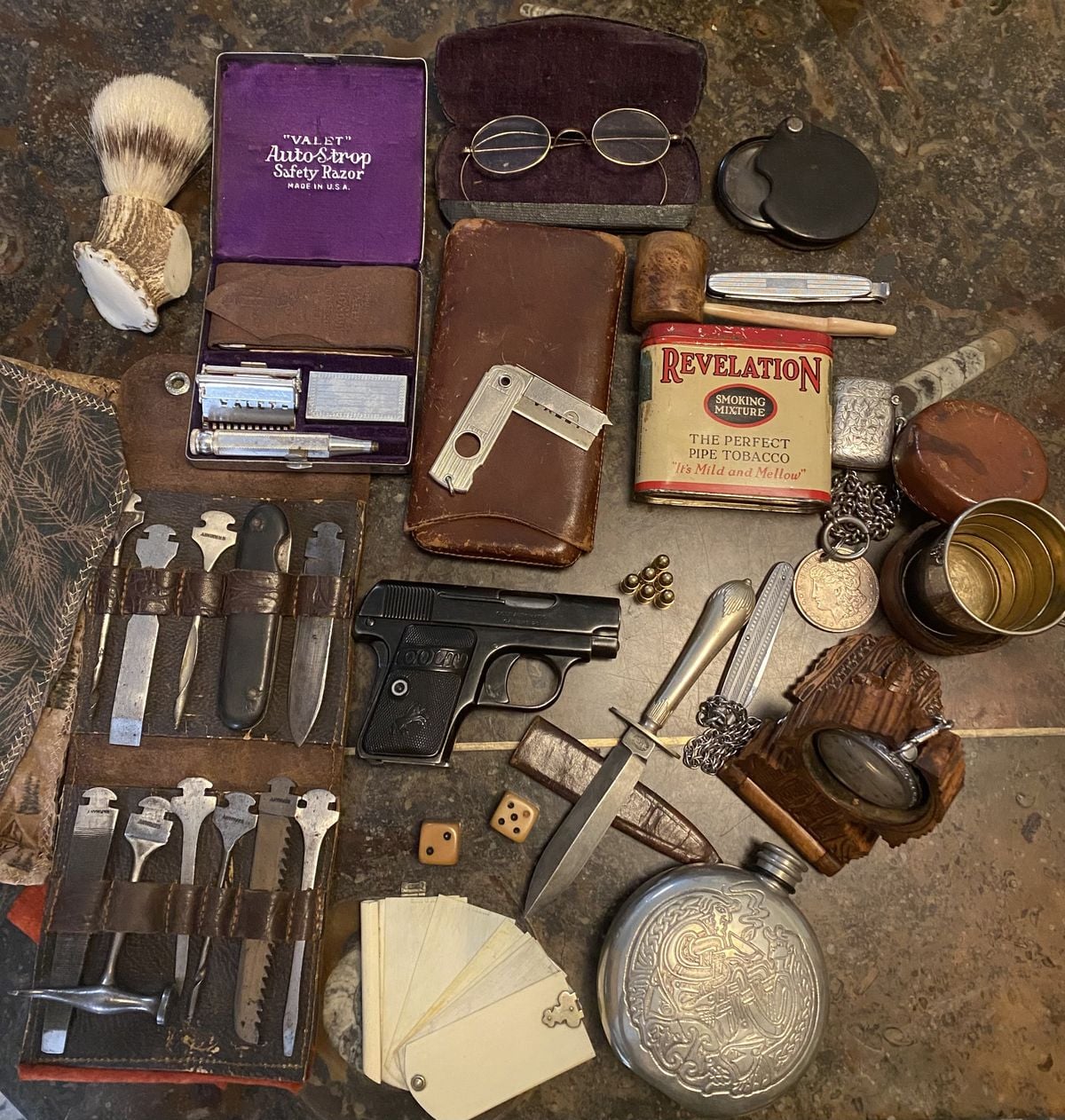 A collection of vintage items on a table including an old pistol, playing cards, safety razor kit, straight razor, shaving brush, tobacco tin, keys, spectacles, knife, notebook, wallet, flask, dice, compass, and pocket watch.