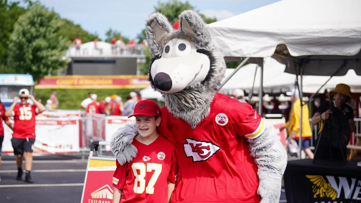 A person in a red Kansas City Chiefs shirt and cap stands next to a large gray wolf mascot, also in a Chiefs jersey, posing for a photo outdoors at a fan event. Tents and other people are visible in the background.