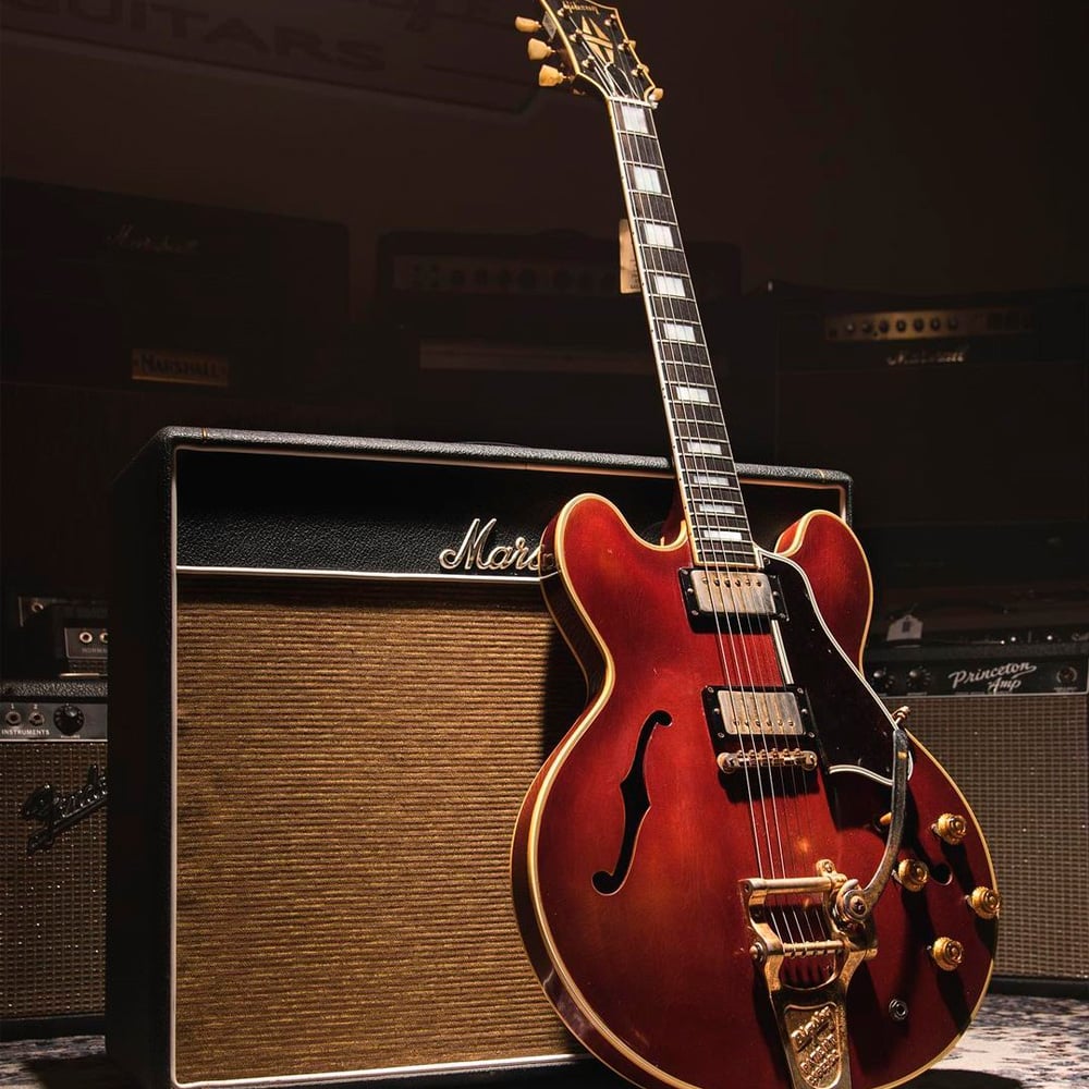A semi-hollow electric guitar with a reddish-brown finish rests against a vintage-style Marshall amplifier, surrounded by other amps in a dimly lit music studio.