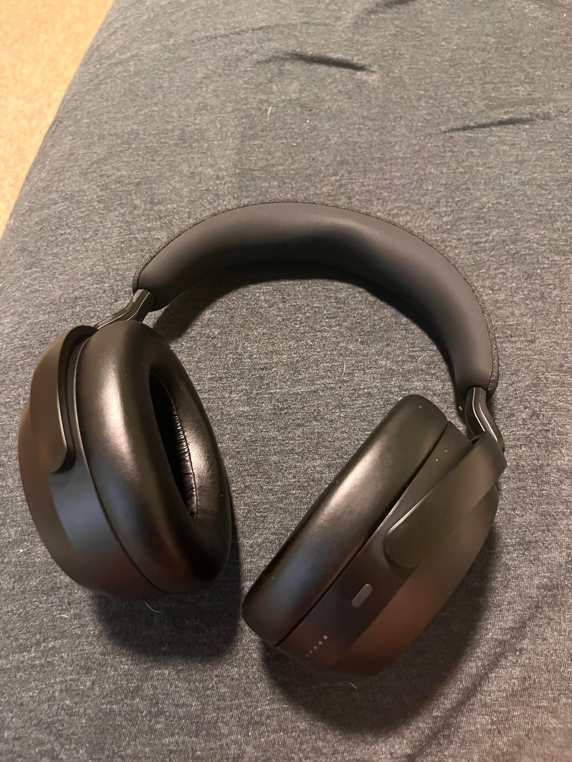A pair of black over-ear wireless headphones is resting on a gray fabric surface. The headphones have cushioned ear pads and a padded headband.