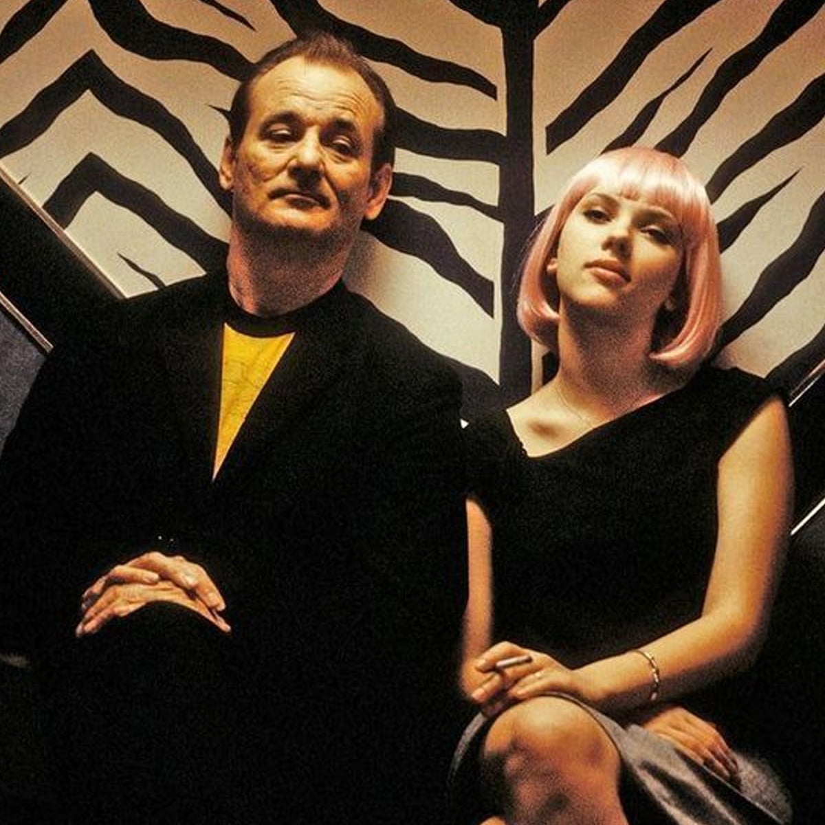 Two people sit side by side on a bench; the man wears a dark suit, and the woman has a short pink wig and a black dress. Black-and-white zebra-patterned decor is visible behind them.