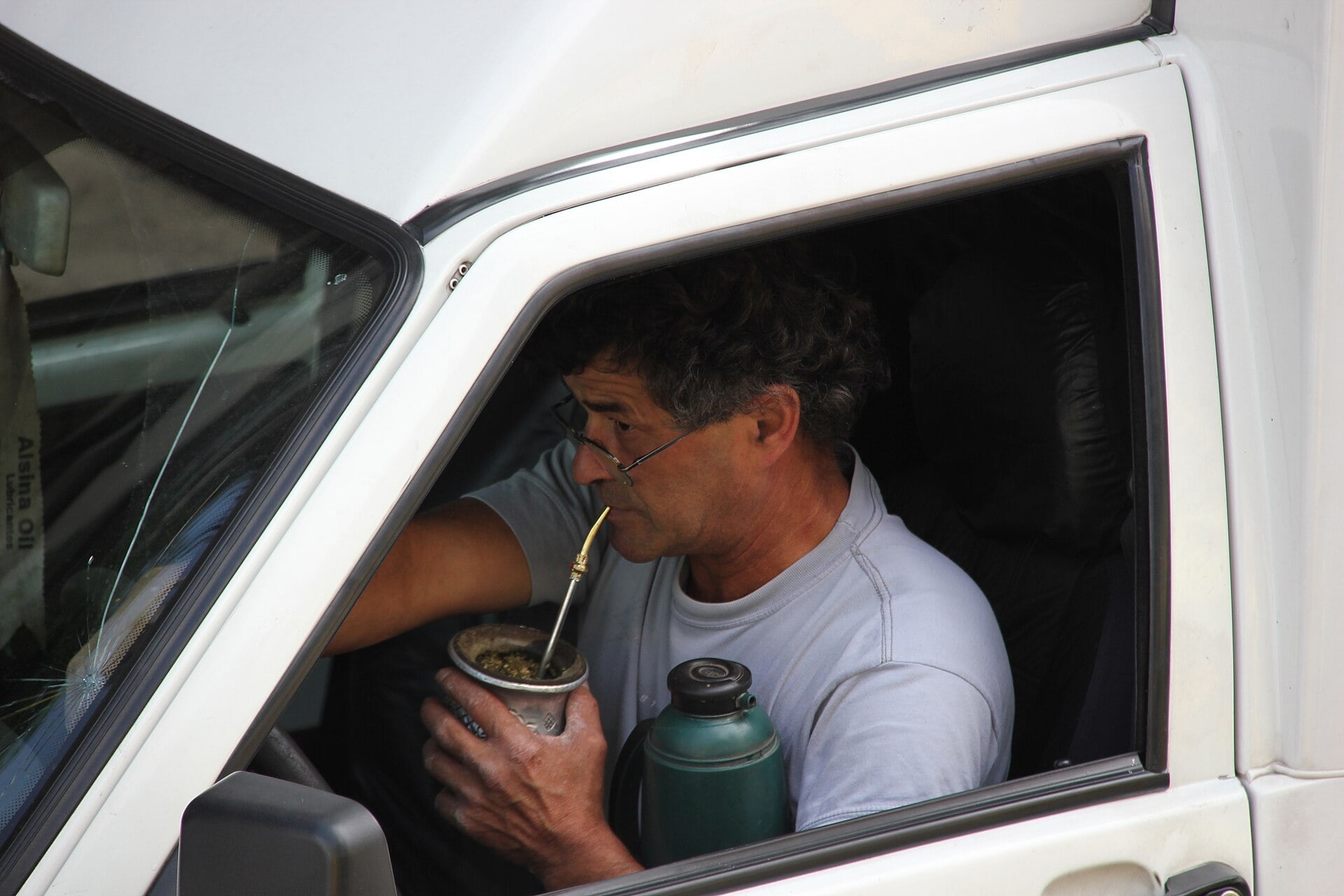 A man sitting in the driver’s seat of a white vehicle drinks yerba mate through a metal straw from a cup, with a green thermos resting beside him.
