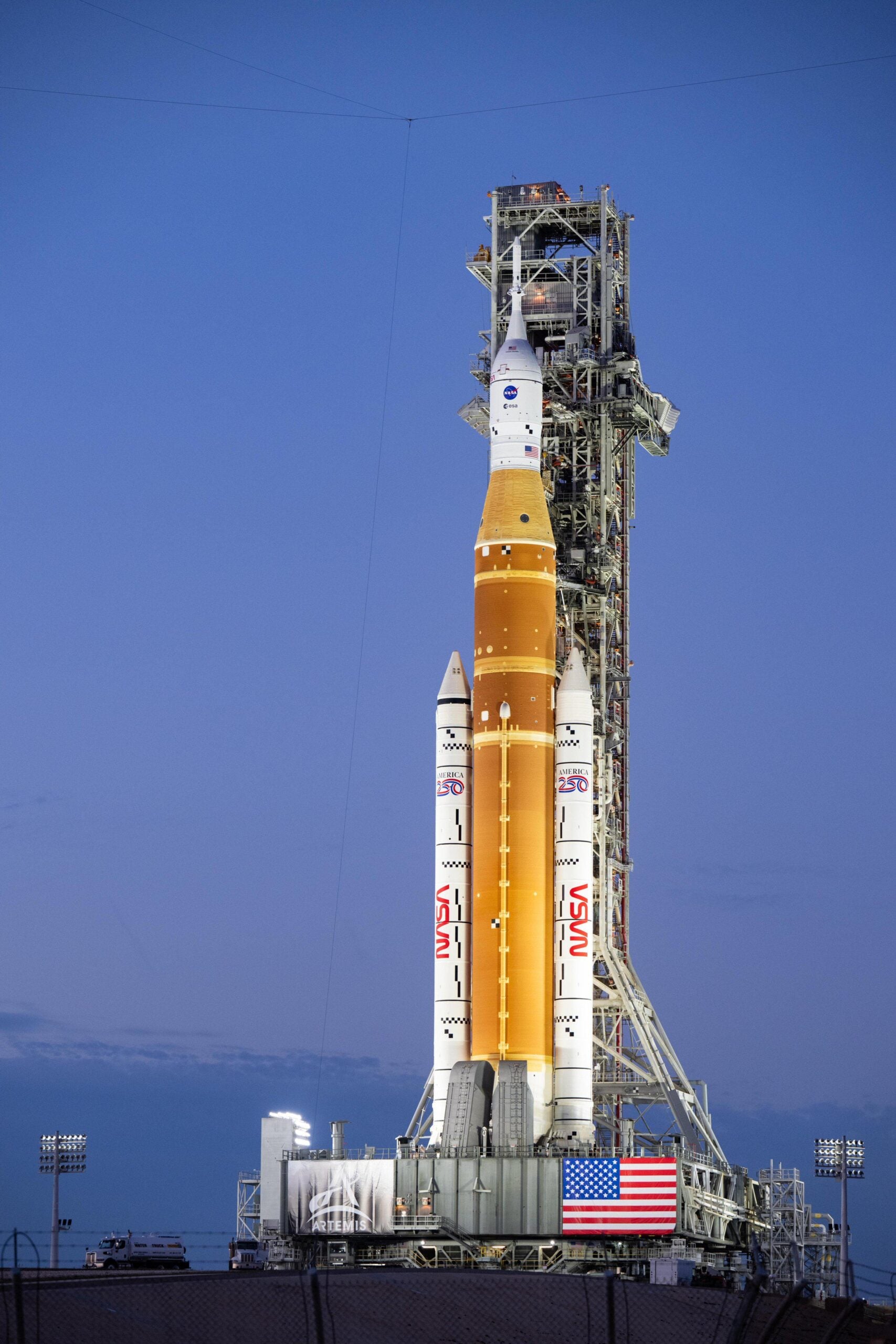 A NASA Space Launch System (SLS) rocket stands upright on a launch pad at dusk, flanked by support structures and lit by floodlights, with an American flag displayed on the platform in the foreground.