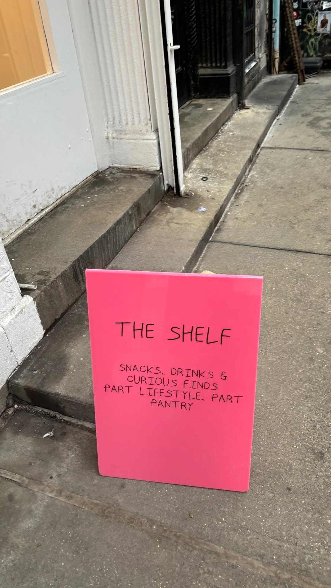 A bright pink sandwich board on a sidewalk reads: "THE SHELF. Snacks, drinks & curious finds. Part lifestyle. Part pantry." It stands near a white building entrance.