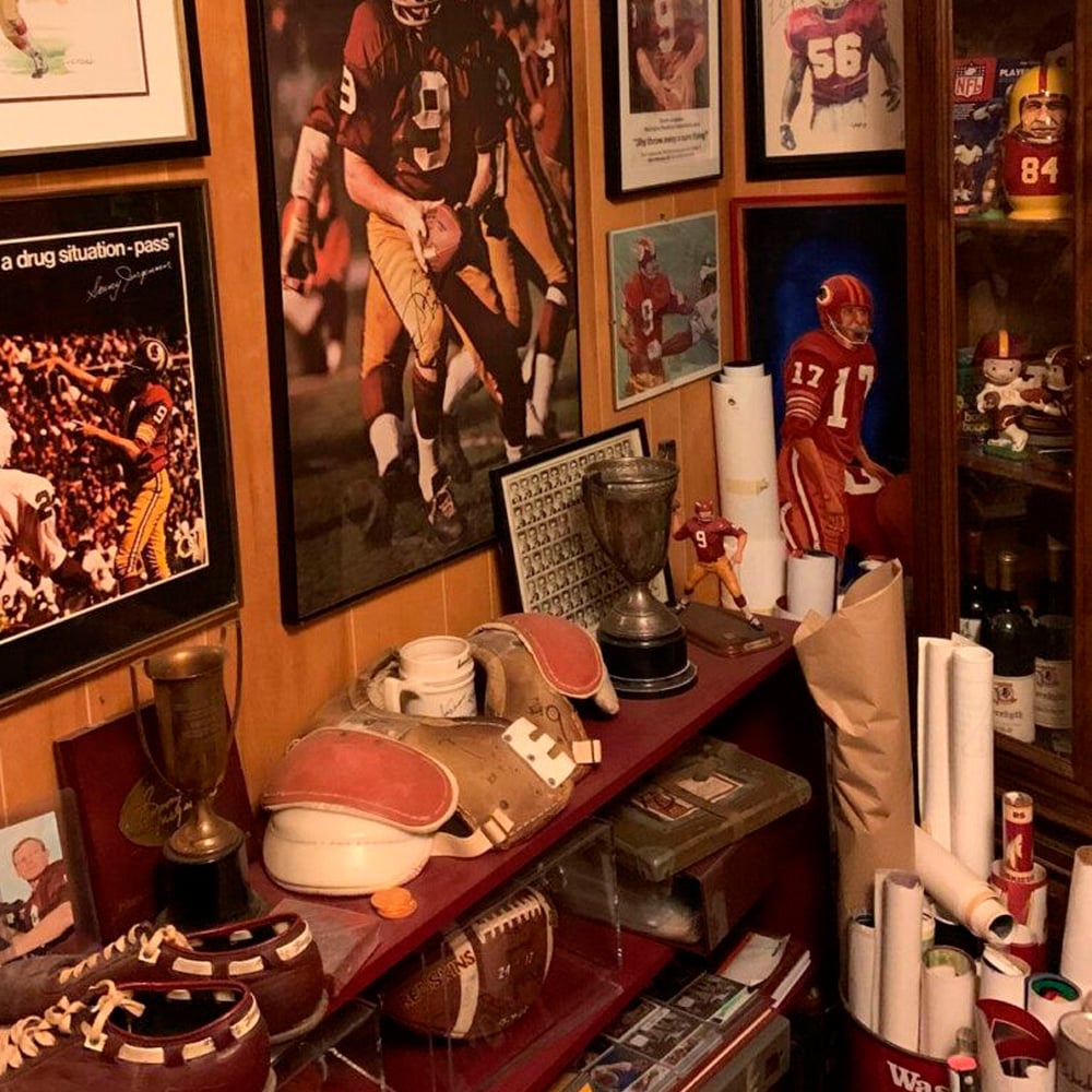 A room filled with football memorabilia, including framed photos and jerseys on the walls, trophies, footballs, old shoulder pads, and helmets on shelves and a cabinet, with posters and rolled-up papers nearby.