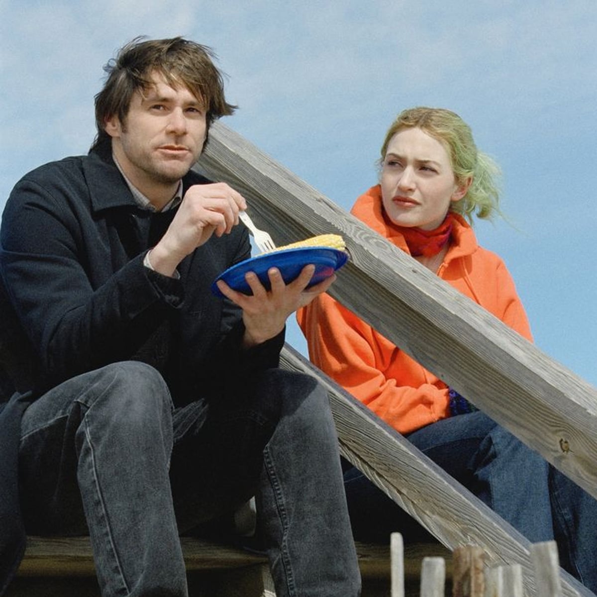 A man holding a plate and eating with a fork sits next to a woman with green hair wearing an orange coat. They are outdoors on wooden steps under a blue sky.