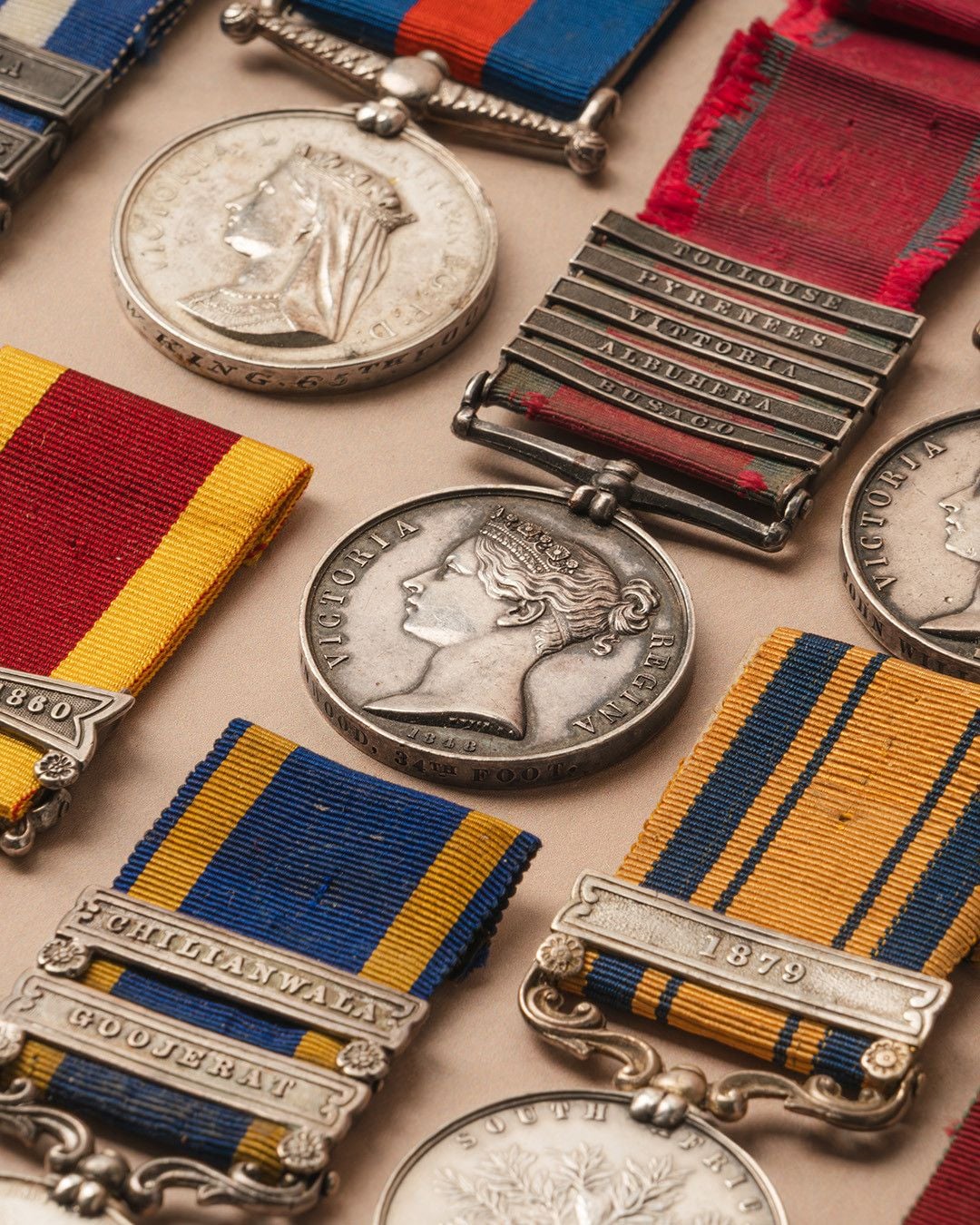 Close-up of several antique British military medals and ribbons, featuring portraits of Queen Victoria and inscribed metal clasps, neatly arranged on a beige surface.