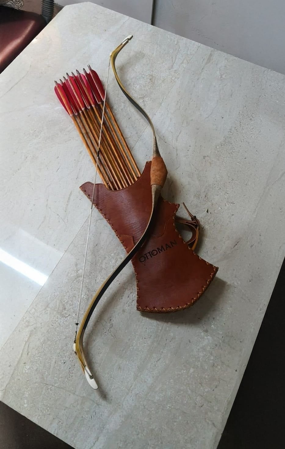A traditional bow and a leather quiver holding several red-feathered arrows are placed on a light-colored marble surface. The quiver is labeled “OTTOMAN.”