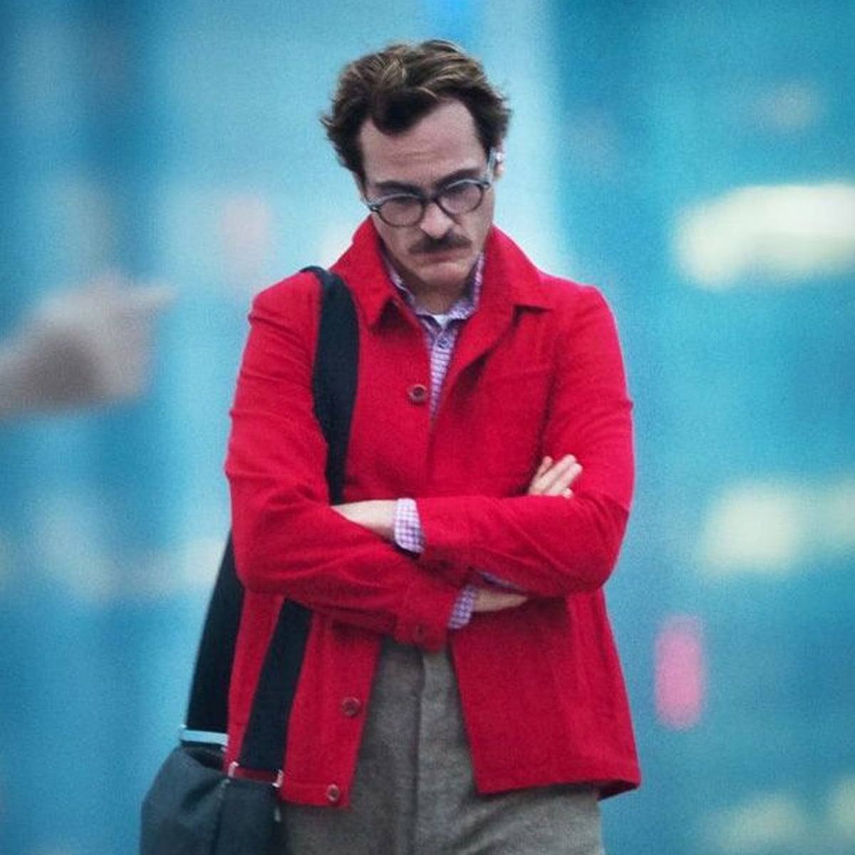 A man wearing glasses and a red jacket stands with his arms crossed, looking down thoughtfully. He has a mustache, carries a shoulder bag, and appears to be outdoors in a city with a blurry blue background.