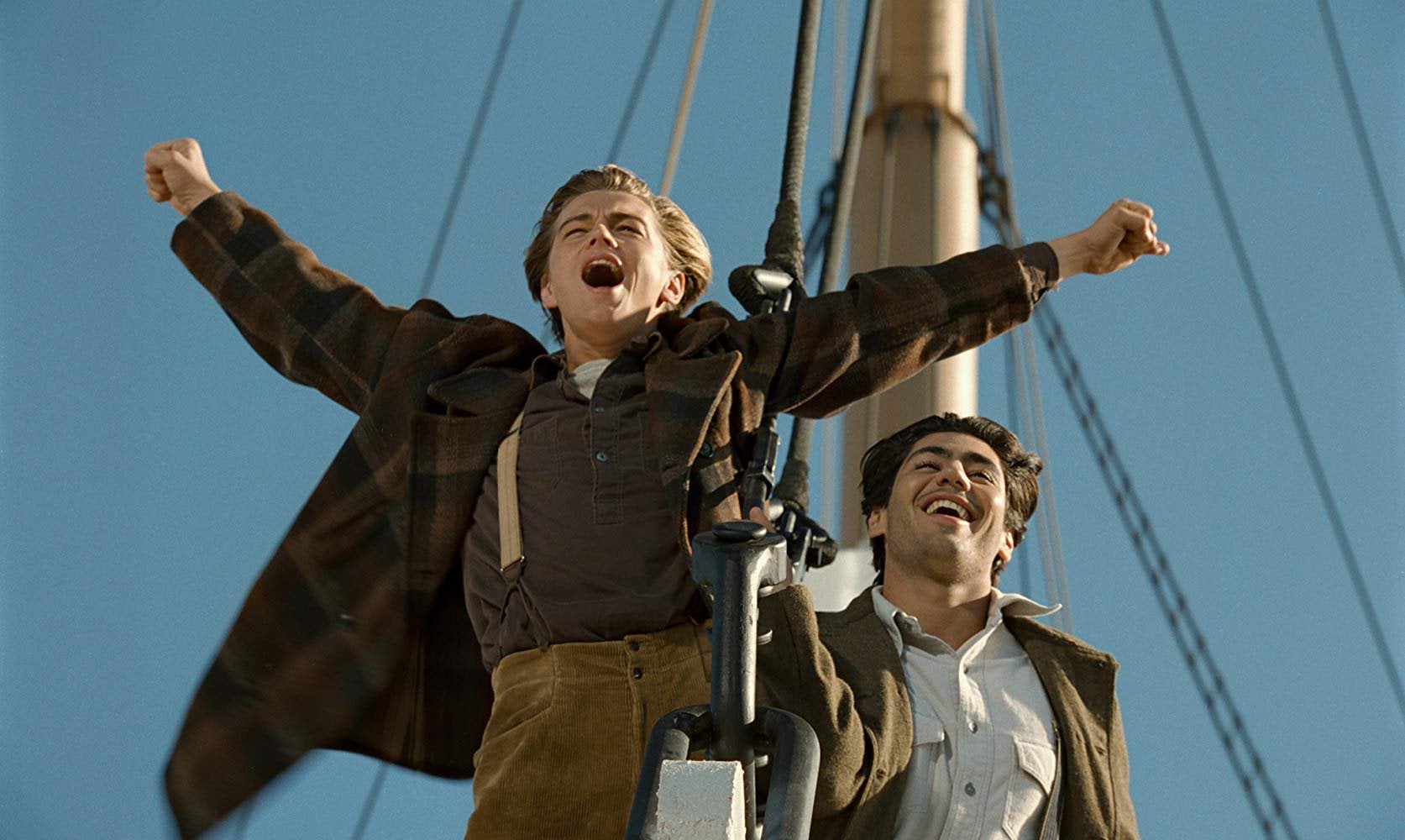 Two men stand at the bow of a ship, one with arms outstretched and an excited expression, the other smiling beside him, with a clear blue sky in the background.