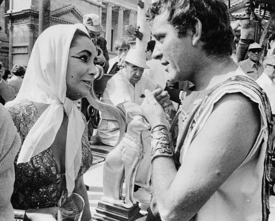 Two actors in historical costumes talk on a movie set. The woman, wearing a headscarf and dramatic eye makeup, drinks from a cat-shaped straw, while the man in ancient attire watches. Crew members and buildings are visible in the background.