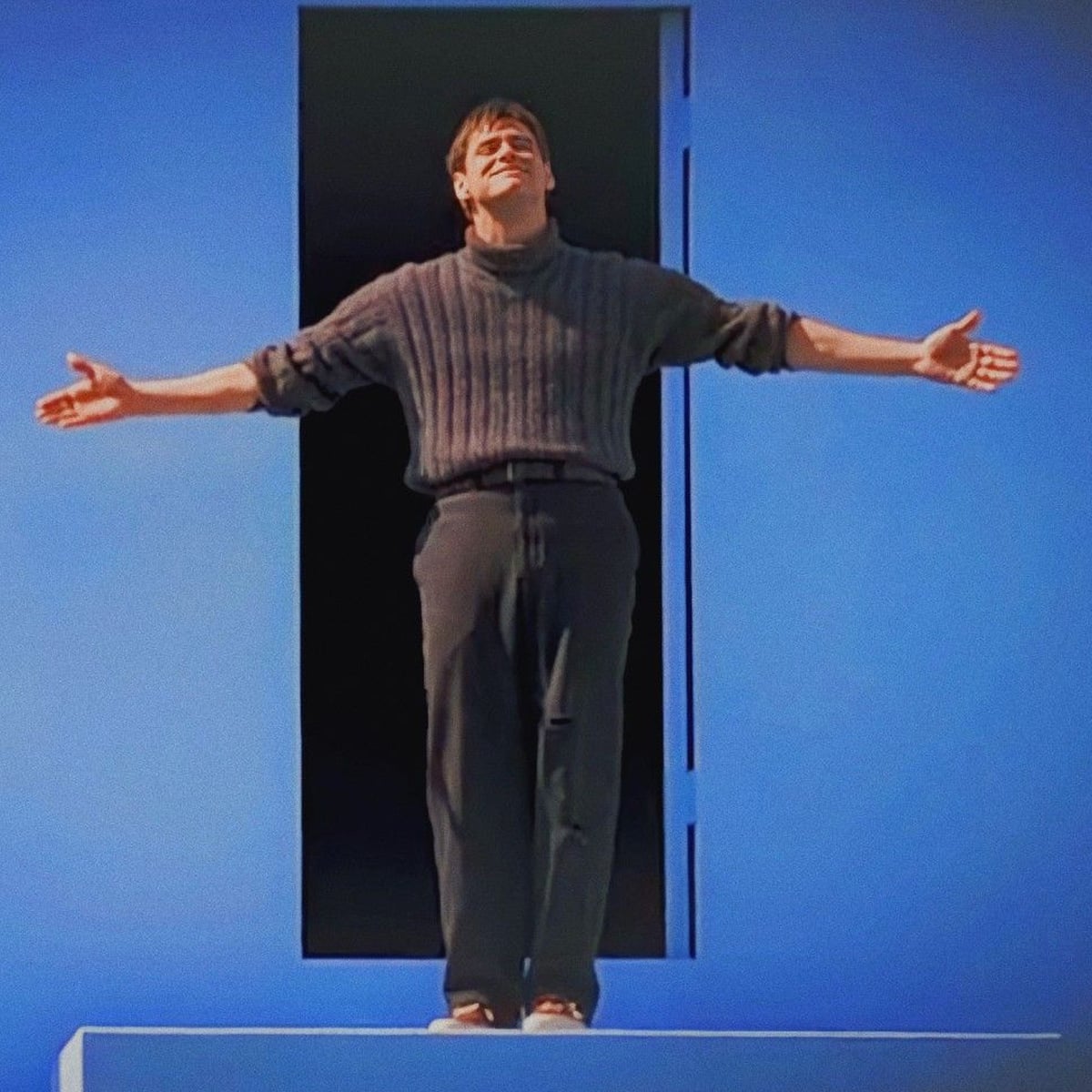 A man stands barefoot on a ledge in front of a large rectangular doorway, arms outstretched, smiling upward. The background is a vibrant, solid blue.