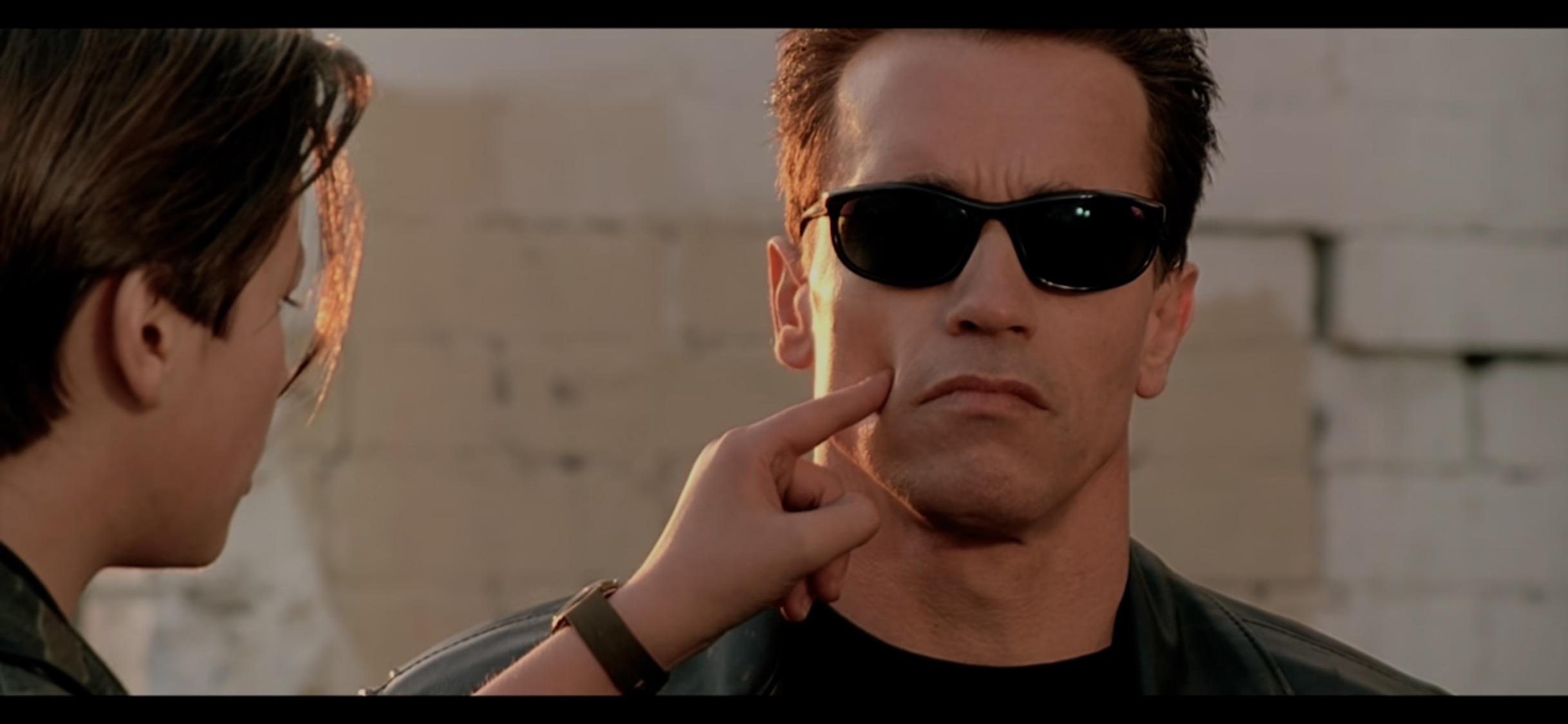 A young person pokes the cheek of a stern-looking man wearing black sunglasses and a leather jacket, who remains expressionless against a light-colored brick wall background.