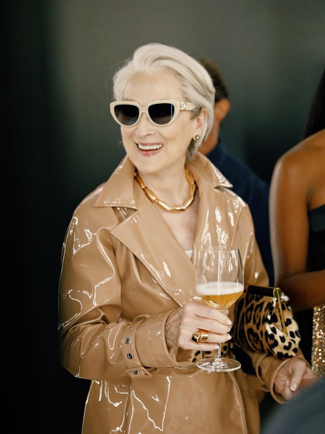 An older woman with short white hair wears a shiny beige trench coat, chunky gold necklace, and cream sunglasses, smiling while holding a glass of white wine. A leopard print bag is seen next to her.