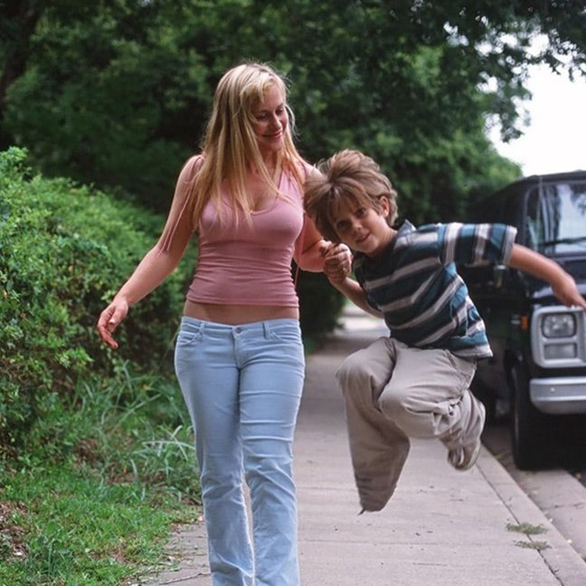 A woman in a pink tank top and light blue pants walks on a sidewalk, holding hands with a boy in a striped shirt who is joyfully jumping. Trees and a parked vehicle are in the background.