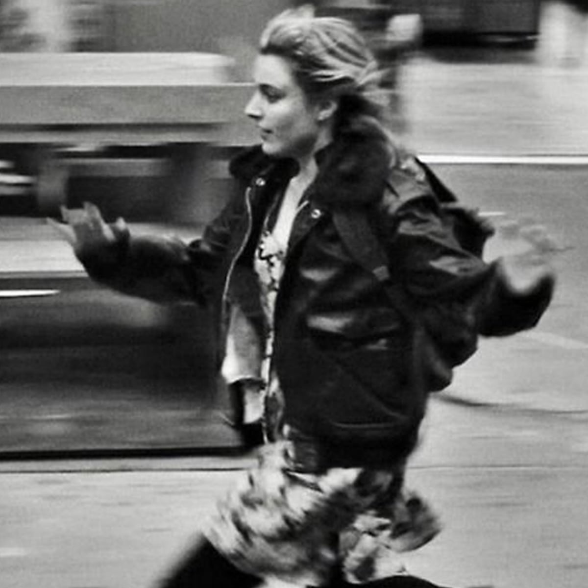 A woman in a patterned dress and dark jacket appears to be running swiftly on a city street, with motion blur creating a sense of movement. The background is out of focus and urban.