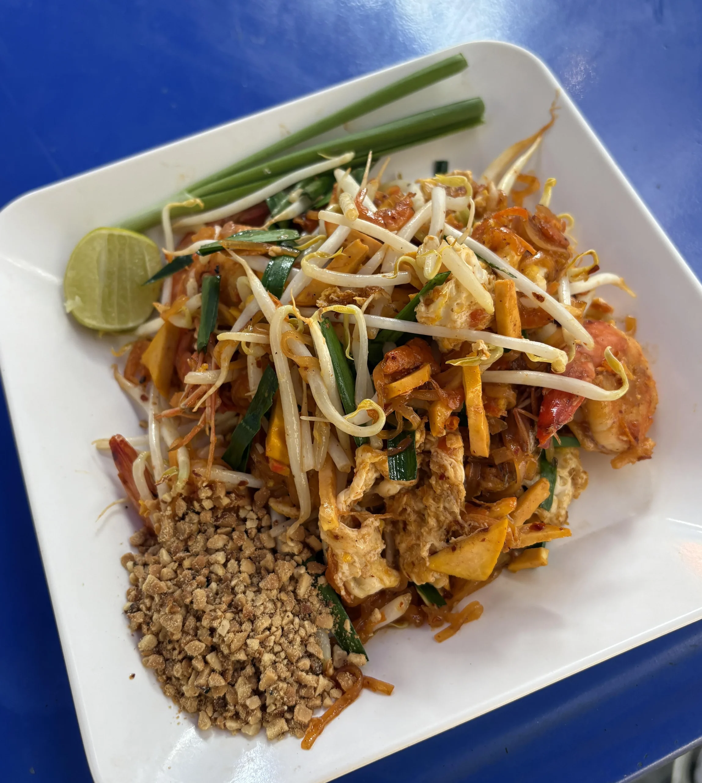 A plate of pad thai topped with bean sprouts and green onions, served with a lime wedge and a pile of crushed peanuts, on a white dish against a blue background.