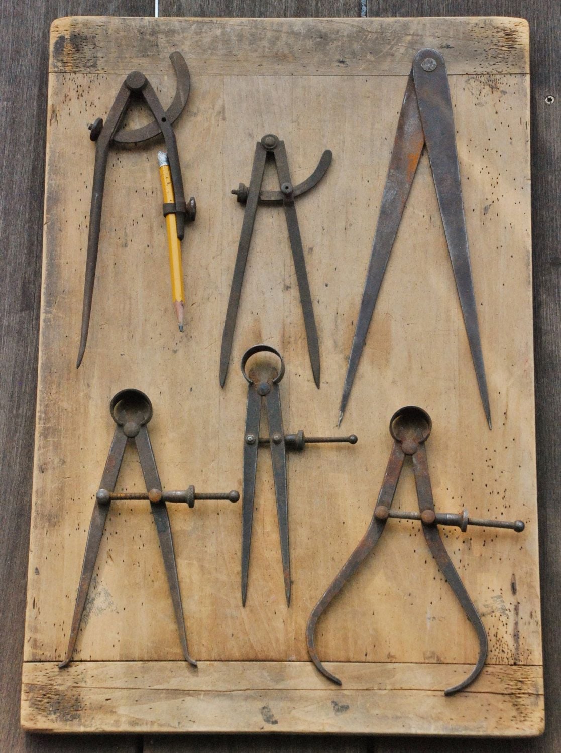 A wooden board displays six vintage metal compasses and dividers, some with adjusting screws. One compass holds a yellow pencil. The tools are arranged neatly against a rustic wooden background.