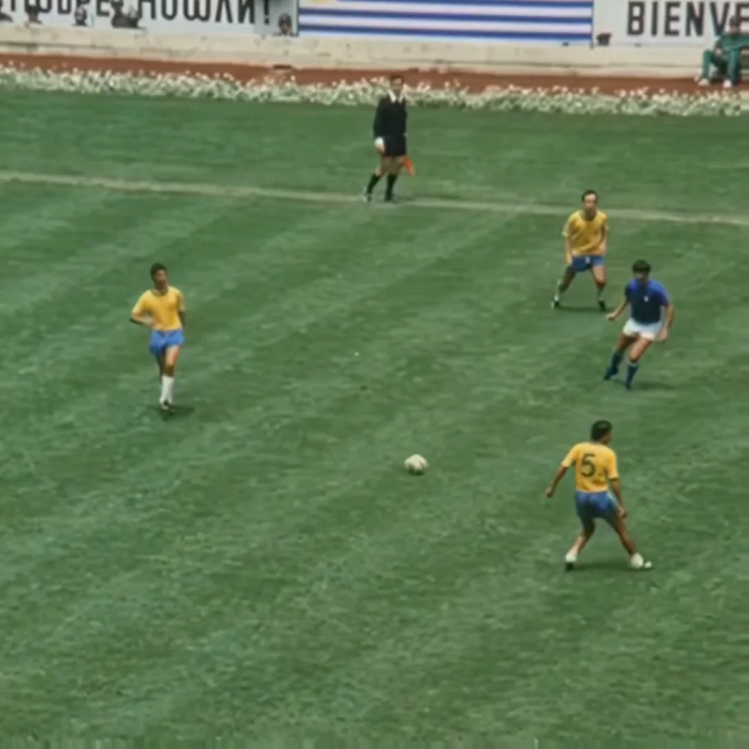 Three soccer players in yellow jerseys and blue shorts face an opponent in blue on a green field, with a referee and a "BIENVENIDOS" sign visible in the background. A soccer ball sits on the grass near the players.